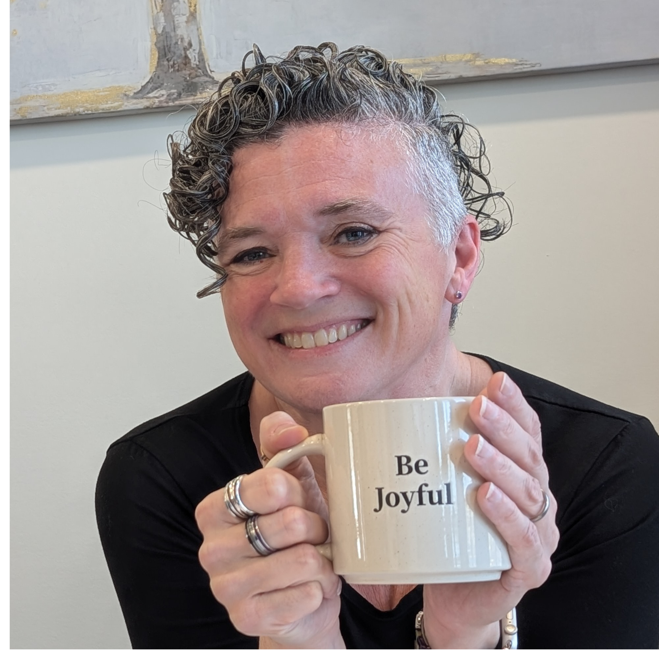 Close-up of a woman (Michelle Renee) with short curly hair smiling, holding a beige mug with black text that reads 'Be Joyful'.