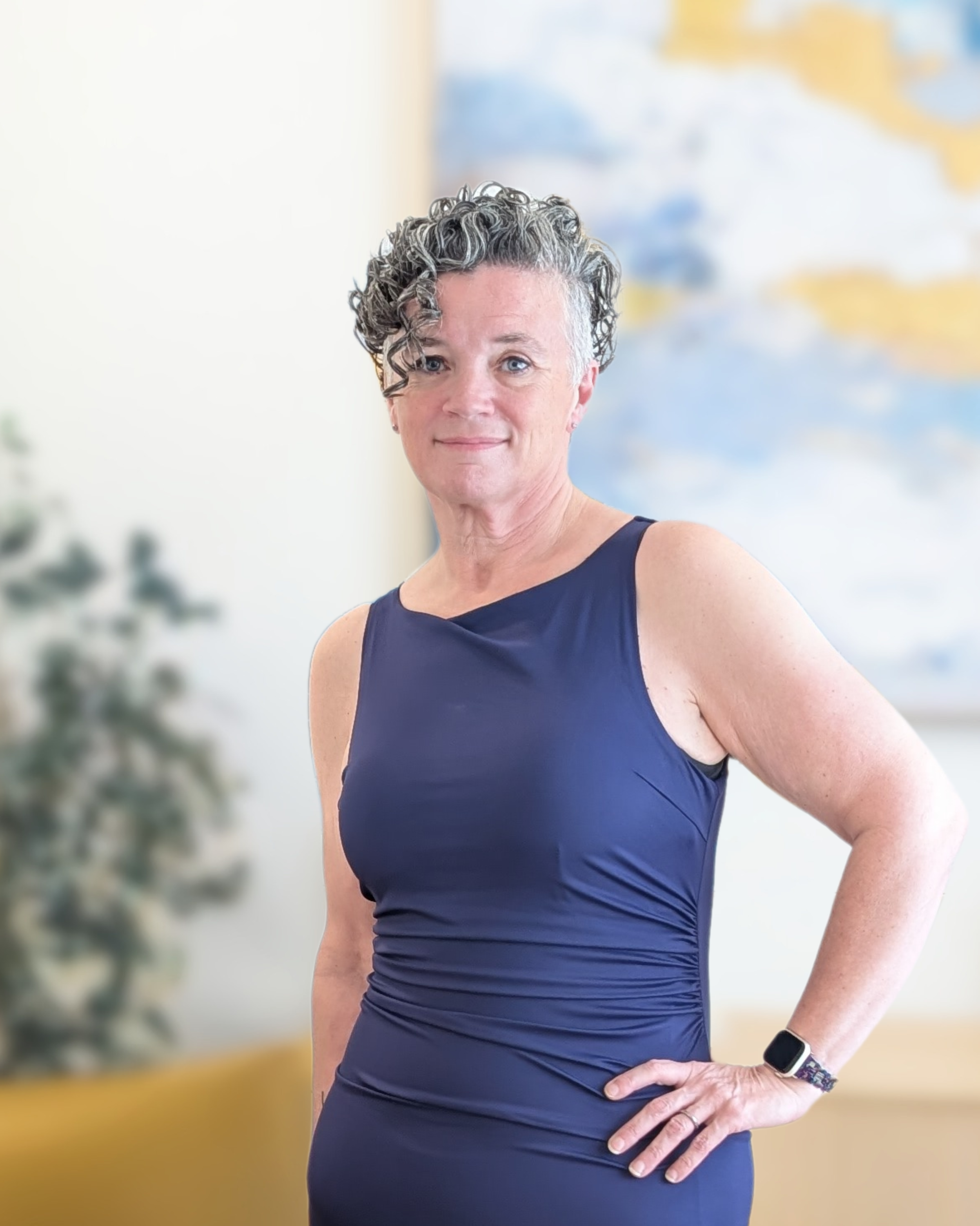 Woman (Michelle Renee) with short curly gray hair wearing a navy sleeveless top and a smartwatch, standing in front of a blurred map and a blurred tree or plant.