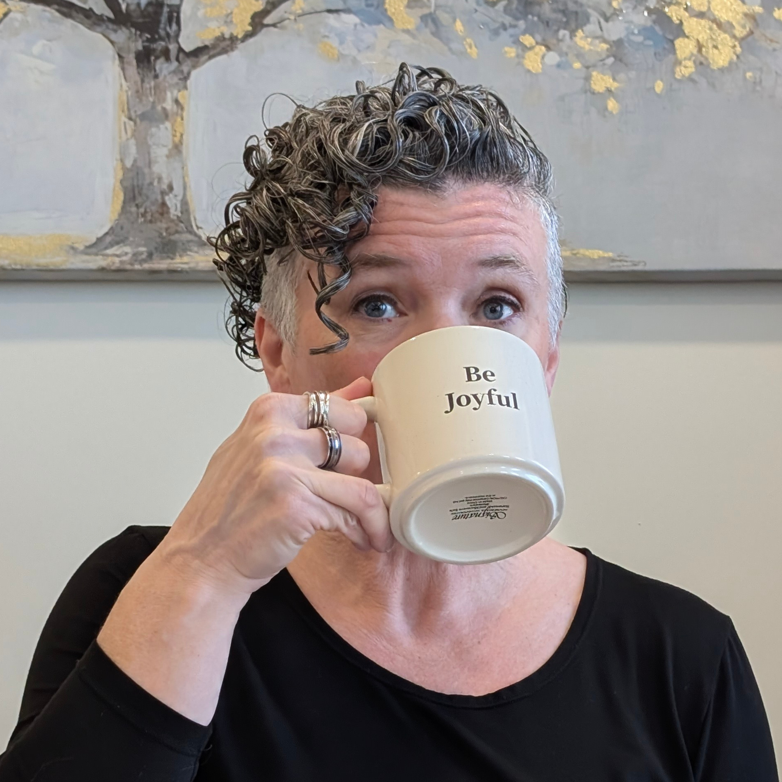 A person (Michelle Renee) with short curly hair is holding a white mug with the words 'Be joyful' written on it covering their face, showing only their eyes and forehead, against a background with a painted tree with yellow leaves.