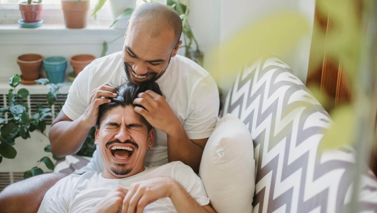 Two men laughing and playing together on a couch, one lying down and the other leaning over him, in a bright room with potted plants.