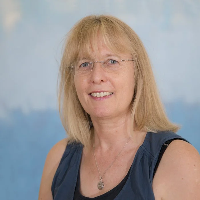 A woman with shoulder-length blonde hair, glasses, and a blue sleeveless top smiling at the camera against a blue background.