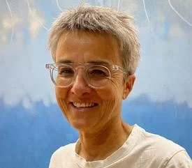 A smiling woman with short gray hair and glasses, wearing a white top, in front of a blue sky background.