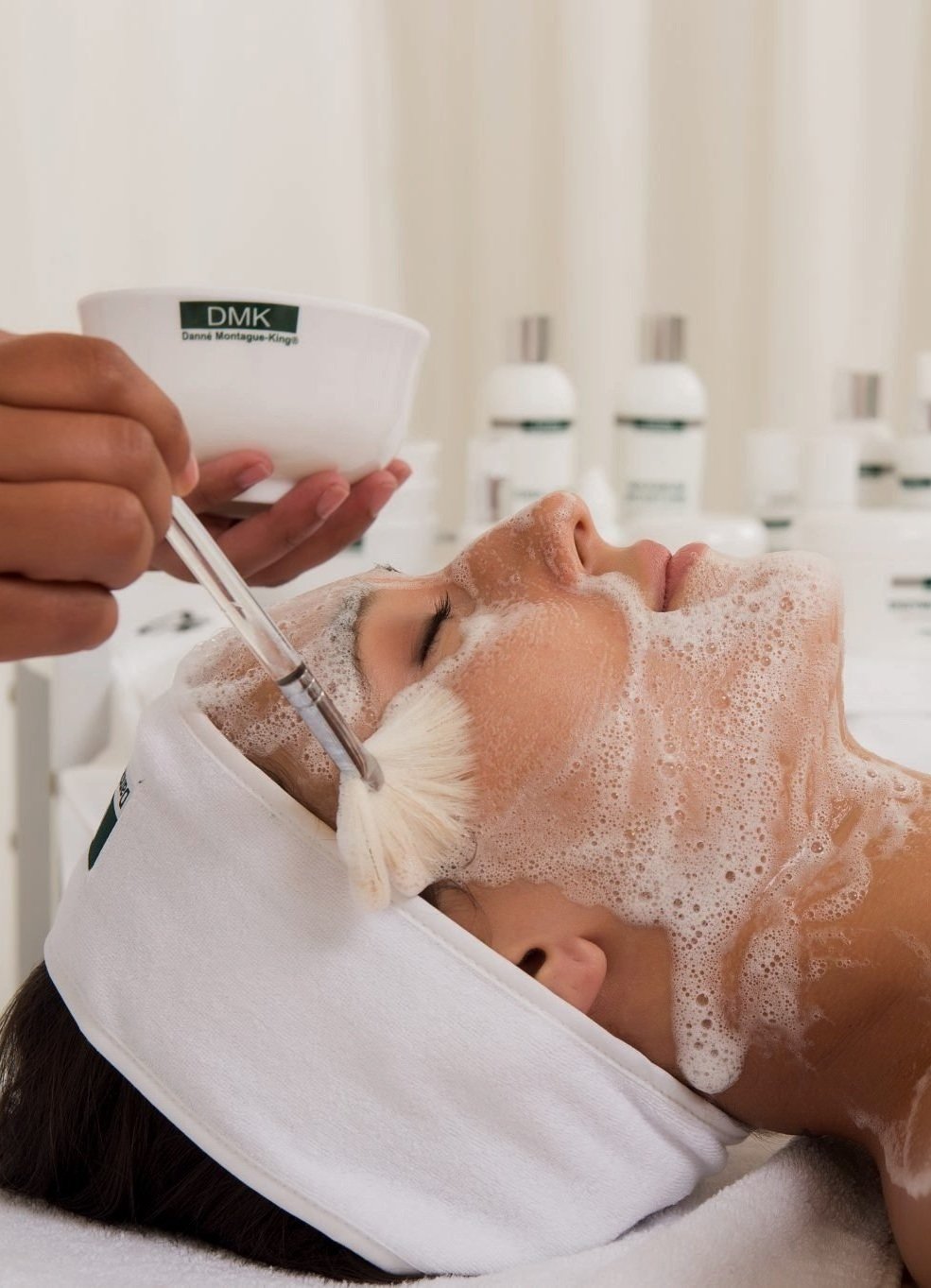 A woman receiving a facial treatment at a spa, lying down with eyes closed, soap lather on her face, and a therapist applying a skincare product using a brush.