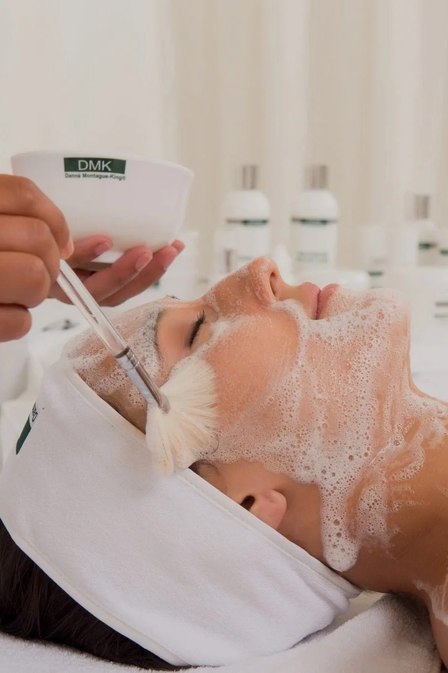 A woman receives a facial treatment, lying down with eyes closed, covered in foam, with a white headband and a technician applying a solution with a brush and a dropper, in a spa or clinic setting.