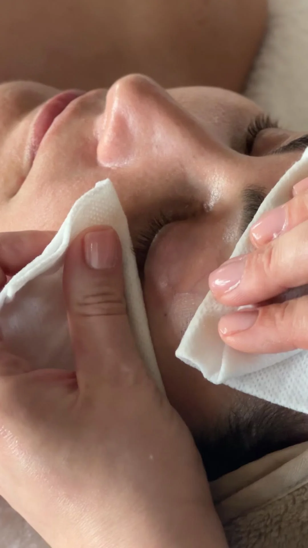 A person receiving a facial treatment, with a close-up of their face and an esthetician wiping their face with a white cloth.