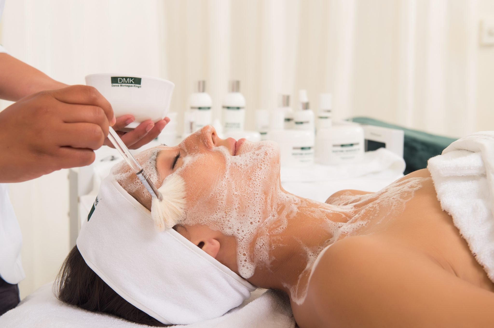 Woman receiving a facial treatment at a spa, with foam and skincare products in the background.
