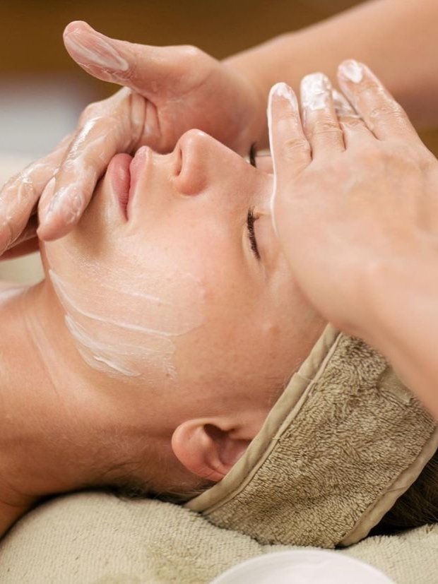 A person receiving a facial massage with their eyes closed, lying down with a towel wrapped around their head, and a light facial mask applied.