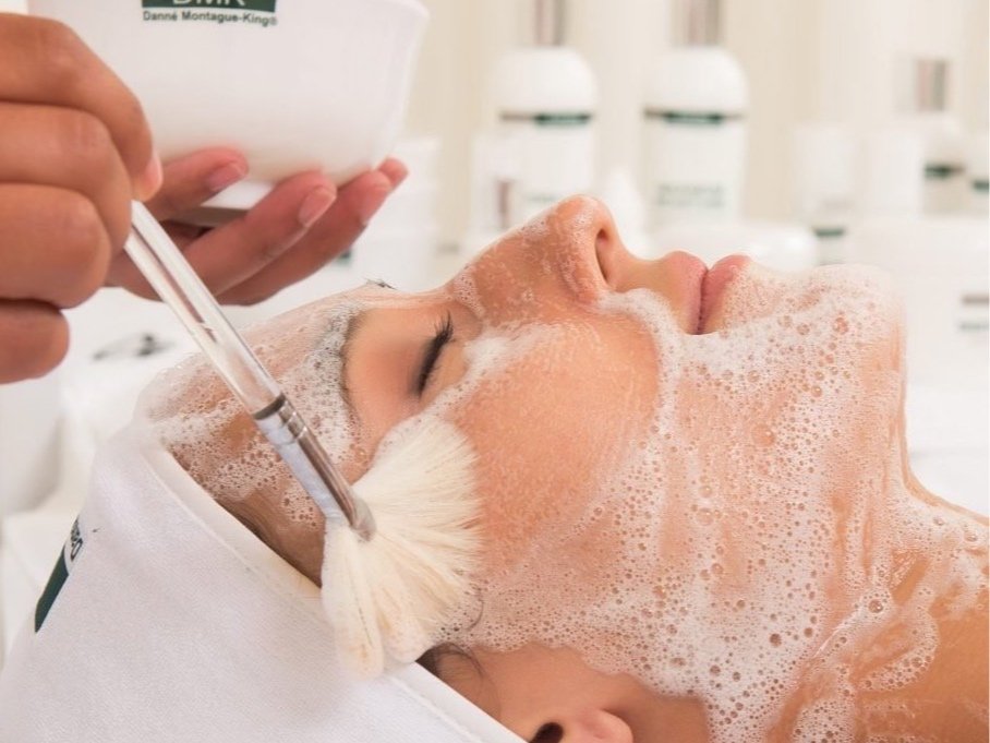 A woman receiving a facial treatment with soap suds on her face in a spa or beauty salon.
