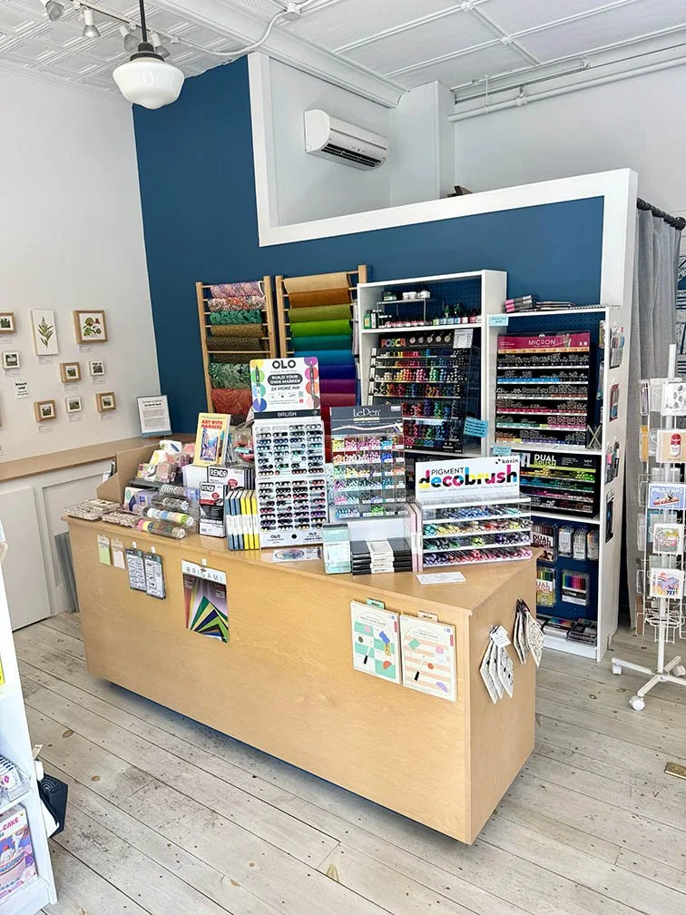 Colorful paper and colored pens merchandised against a blue wall with a large table in front - June refresh 2025