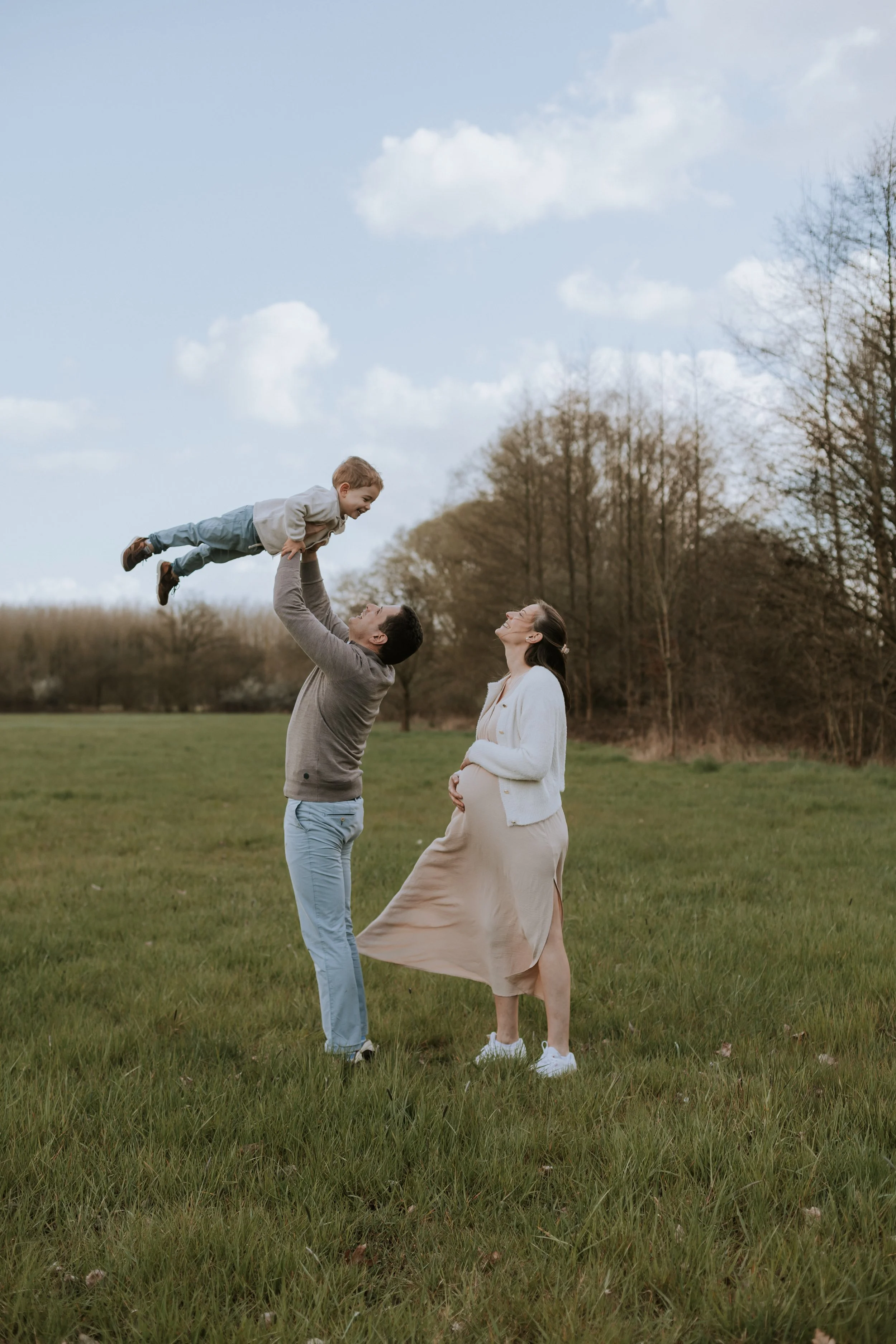 Papa tilt zoontje hoog in de lucht terwijl mama naar ken kijkt en over haar zwangere buik wrijft.