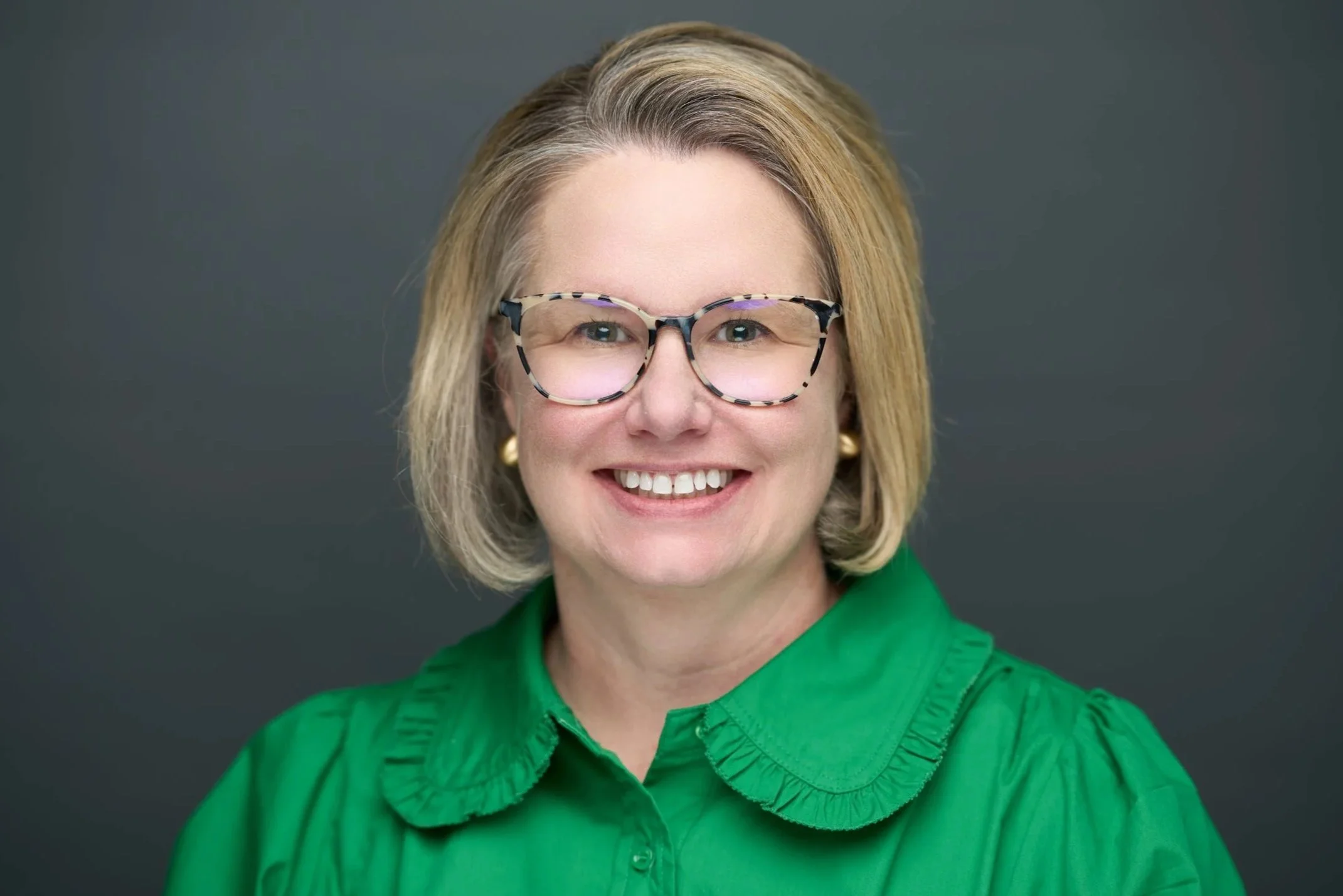 Person smiling, wearing glasses, a black blazer, and a zebra-patterned blouse, against a gray background.
