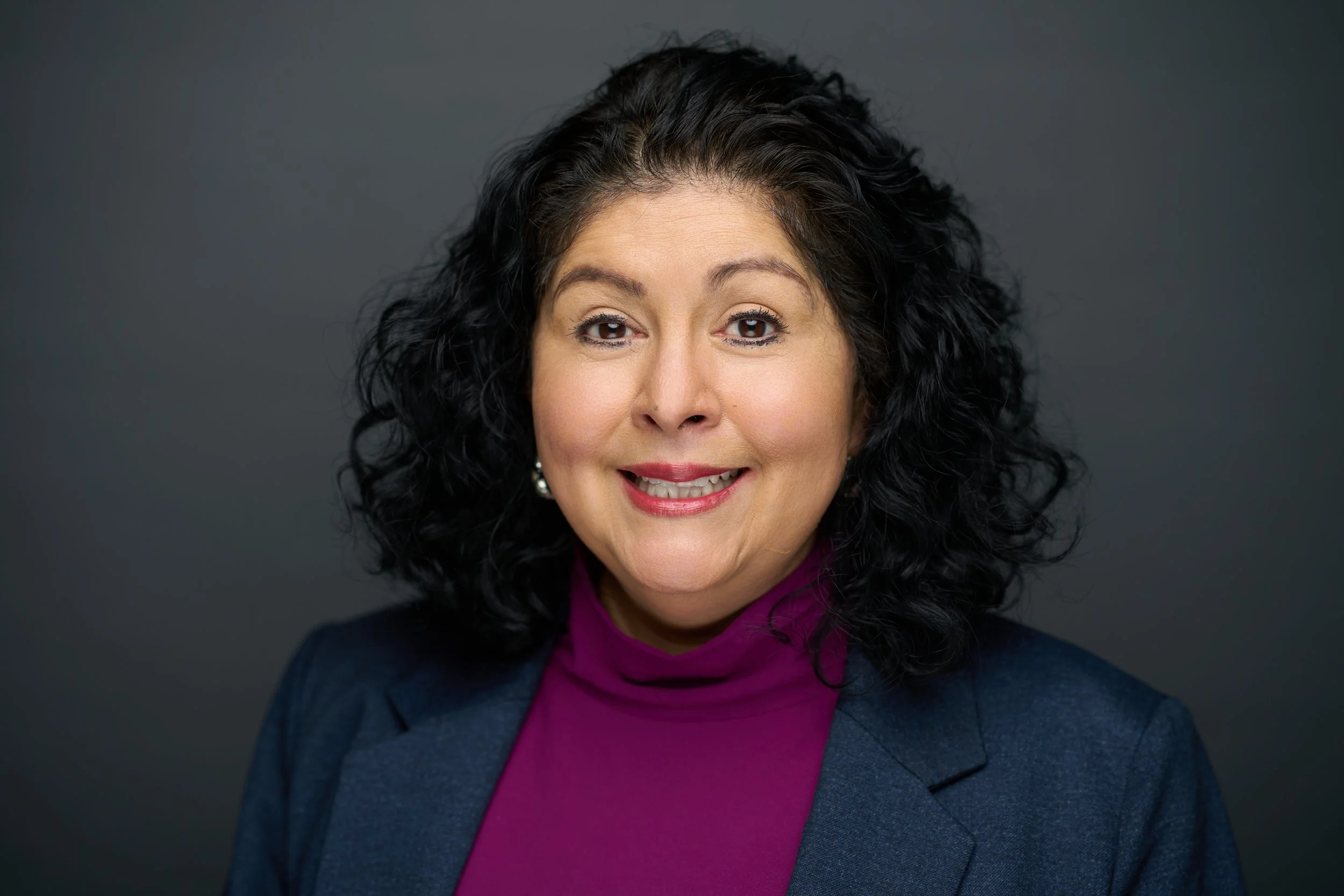 Smiling woman with curly black hair wearing a purple blouse against a gray background.
