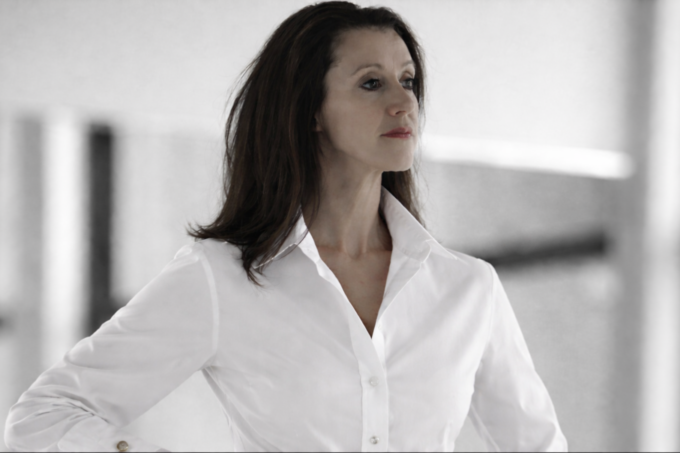Helena Collins with dark brown hair wearing a white button-up shirt, standing in a modern, bright indoor space.