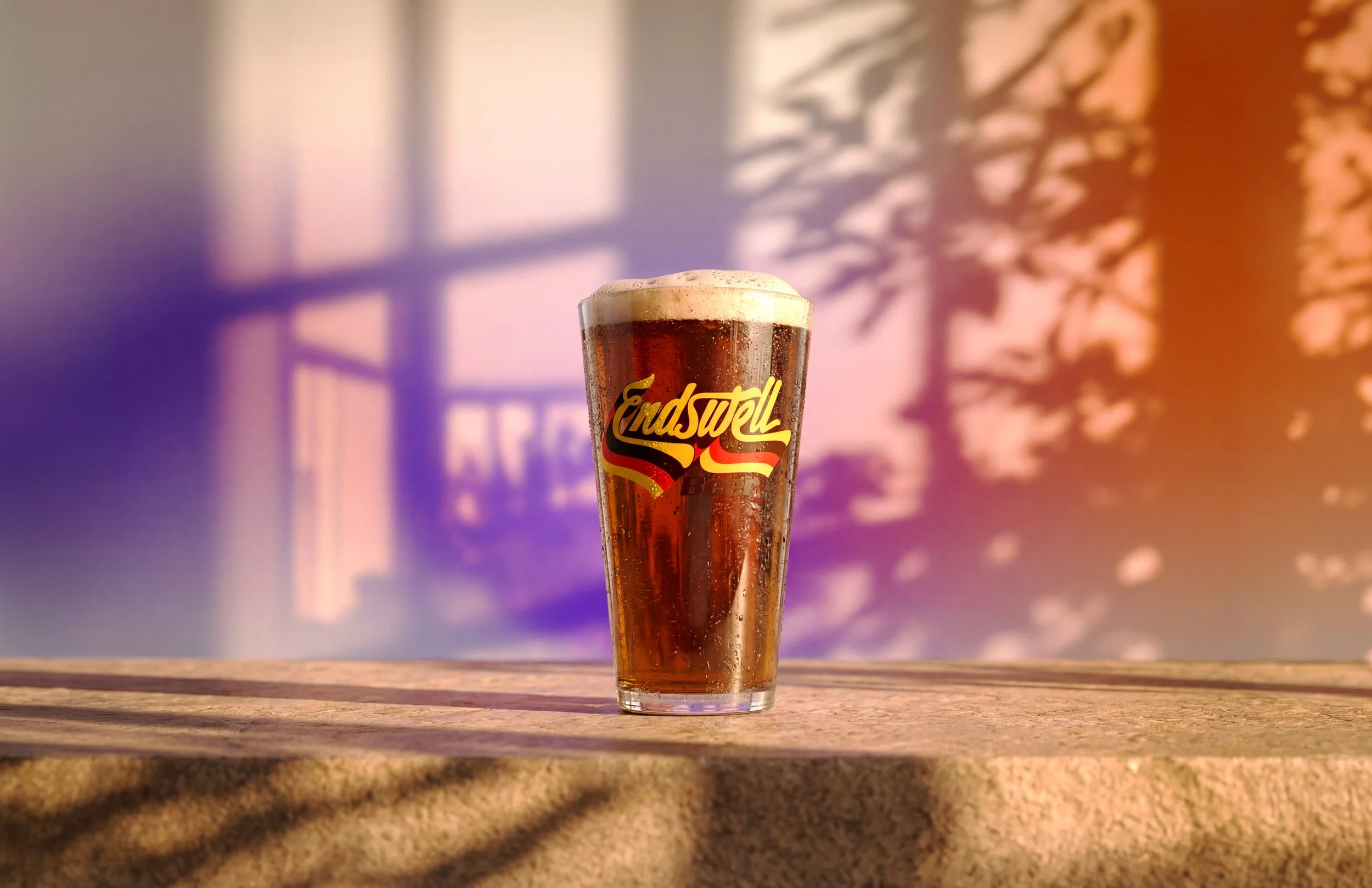 A pint glass of amber beer with foam on top sitting on a wooden surface. The glass has 'Endswell Beer' written on it. In the background, there are blurred window shadows and trees, with sunlight creating a warm, colourful gradient.