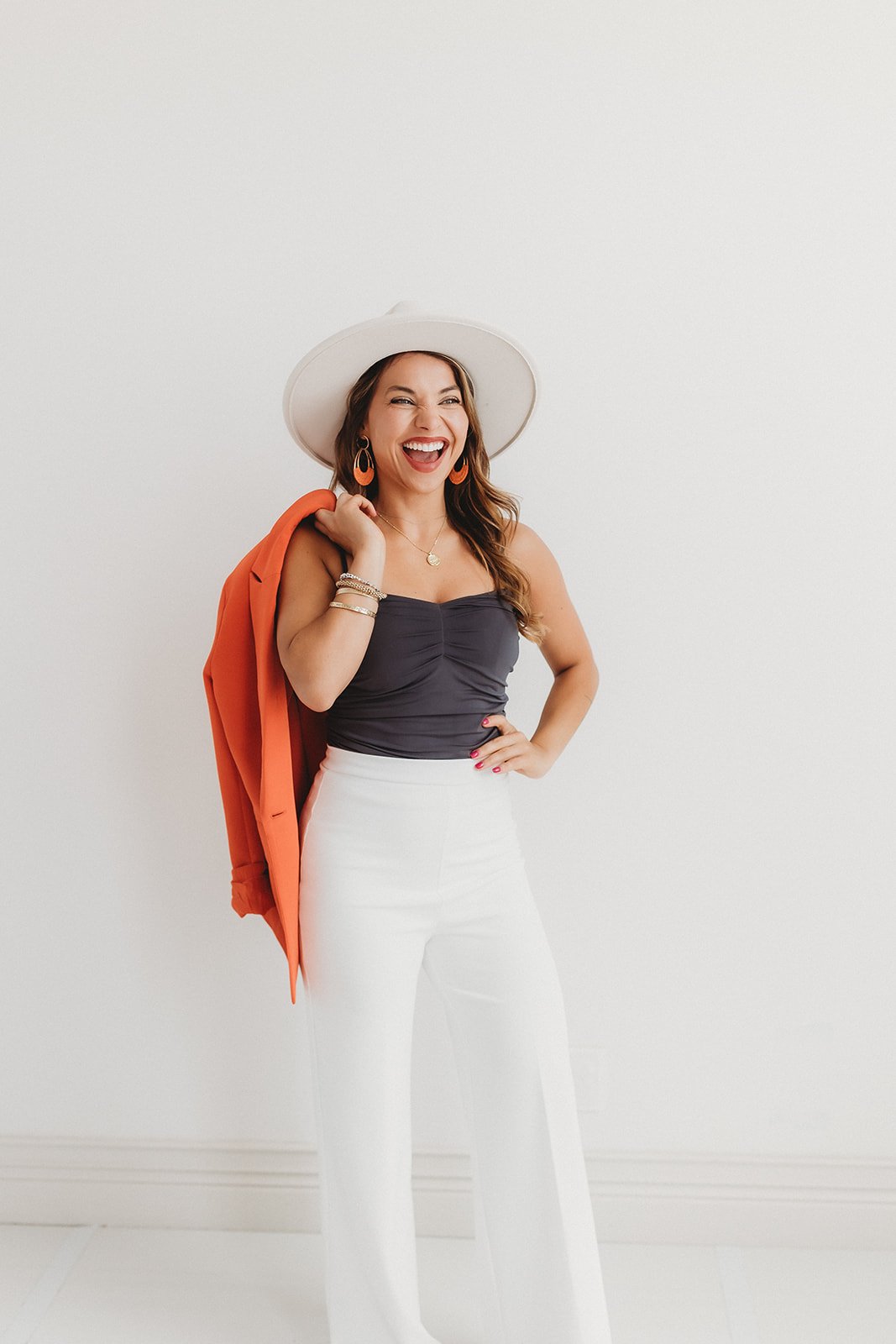 A woman with long brown hair, wearing a wide-brimmed white hat, black strapless top, white high-waisted pants, holding an orange jacket over her shoulder, smiling and posing in front of a plain white wall.