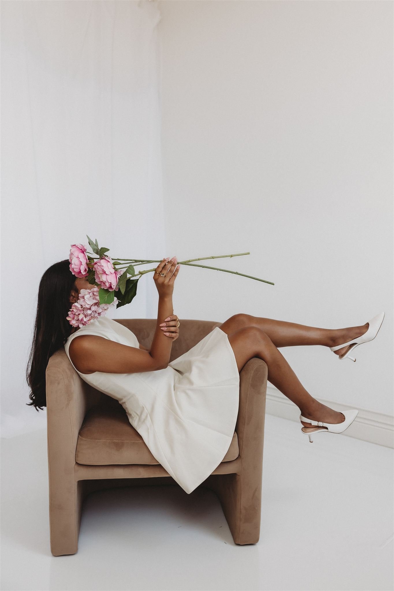A woman lying on a beige armchair, holding pink flowers over her face, wearing a white dress and white high heels, against a white background.