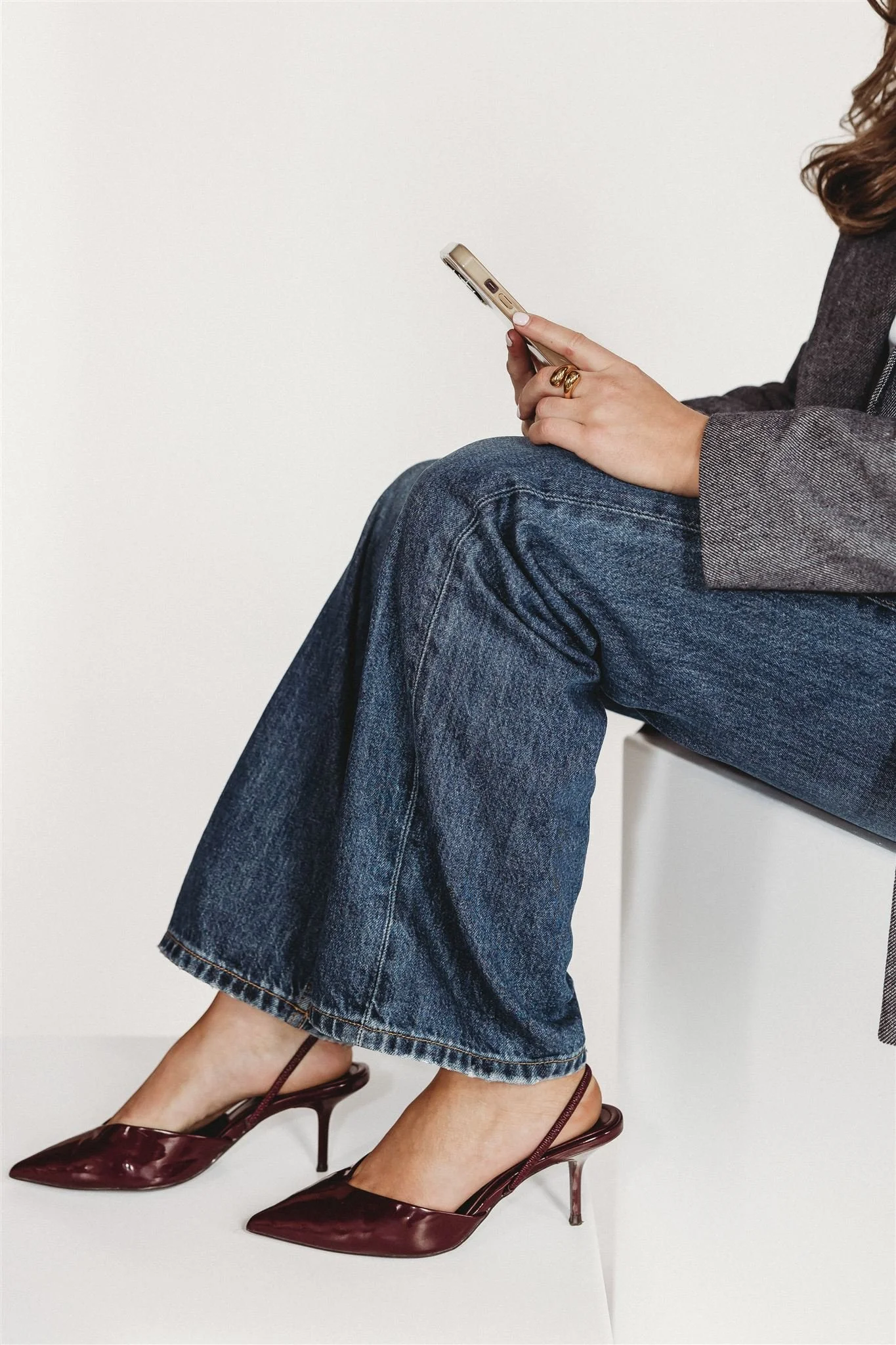 Person sitting on a white surface, wearing blue jeans, a gray blazer, and burgundy high-heeled shoes, using a smartphone.