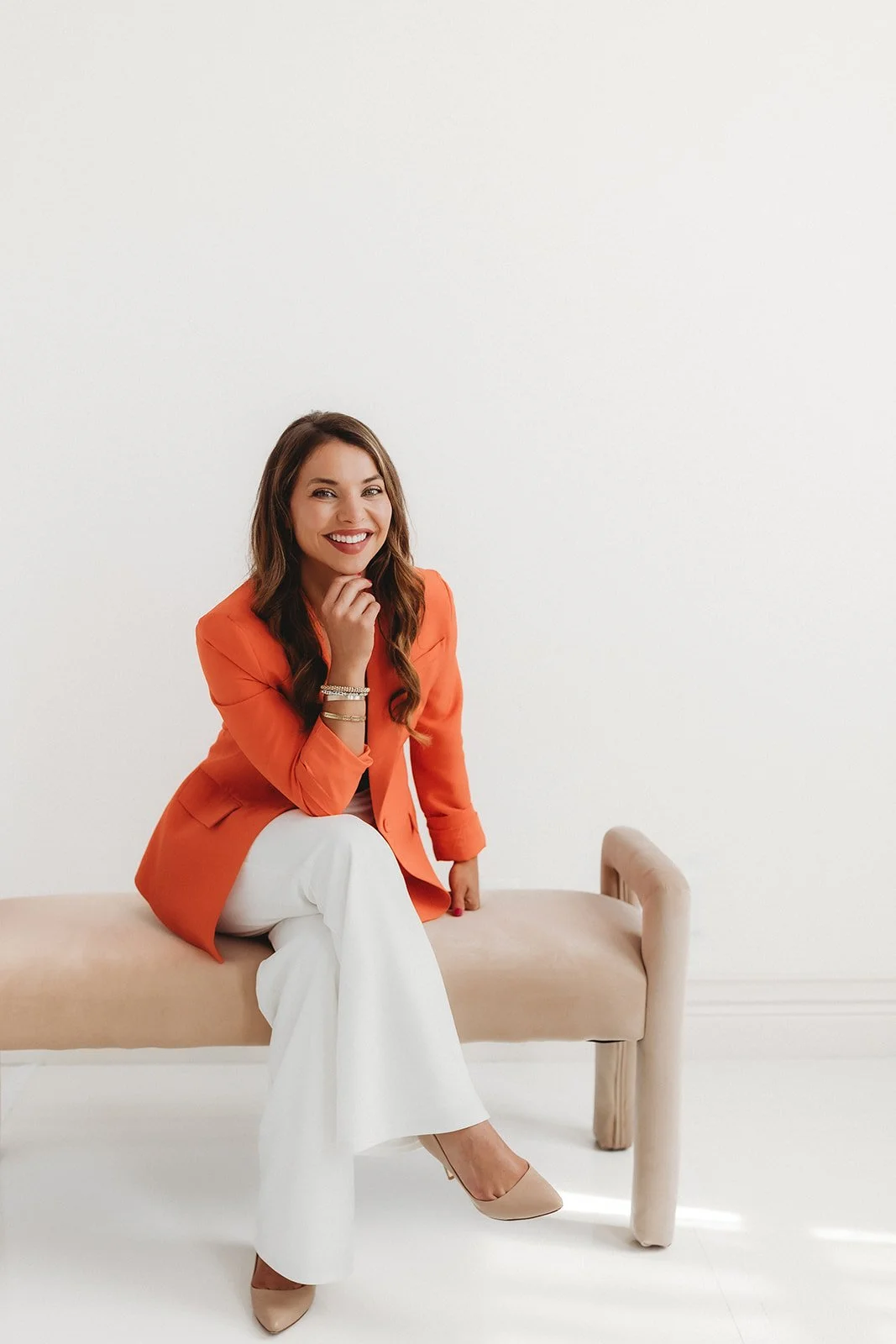 Woman sitting on a bench, wearing an orange blazer and white pants, smiling, in a minimalist white room.