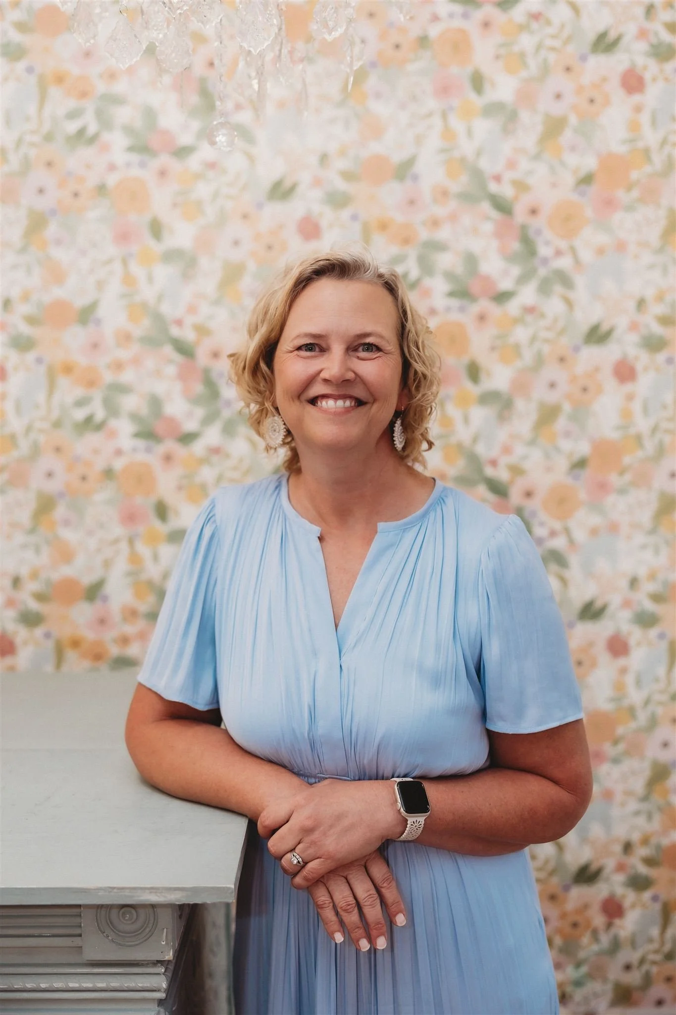A smiling woman with curly blonde hair wearing a light blue pleated dress, standing with her hands clasped in front of her, in front of a floral patterned background.