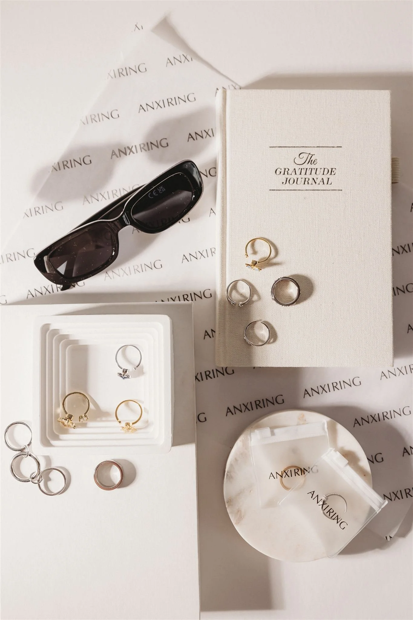 A flat lay of jewelry, sunglasses, and a gratitude journal on a white surface. The jewelry includes rings and earrings, some in gold, silver, and rose gold. The sunglasses are black with a thick frame. The gratitude journal is white with black text. There are pieces of branded paper or tissue with 'ANXIRING' printed on them.