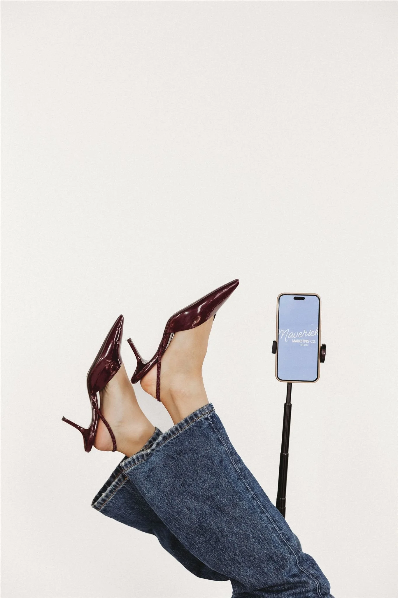 Person's legs raised with polished burgundy high heels and a smartphone on a selfie stick against a plain white background.
