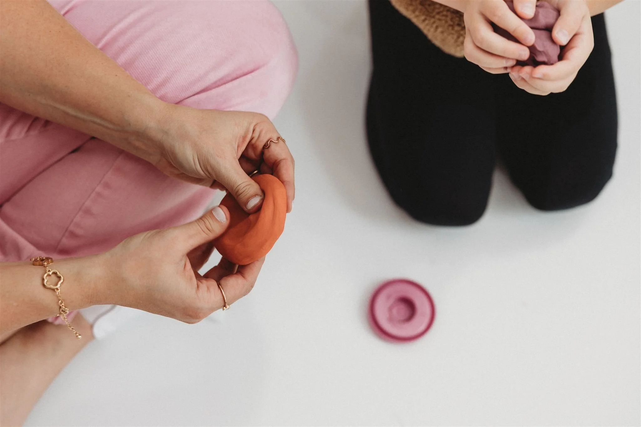 Overhead view of two people sitting across from each other, each kneading colored play dough, with a flattened pink piece on the white floor between them.