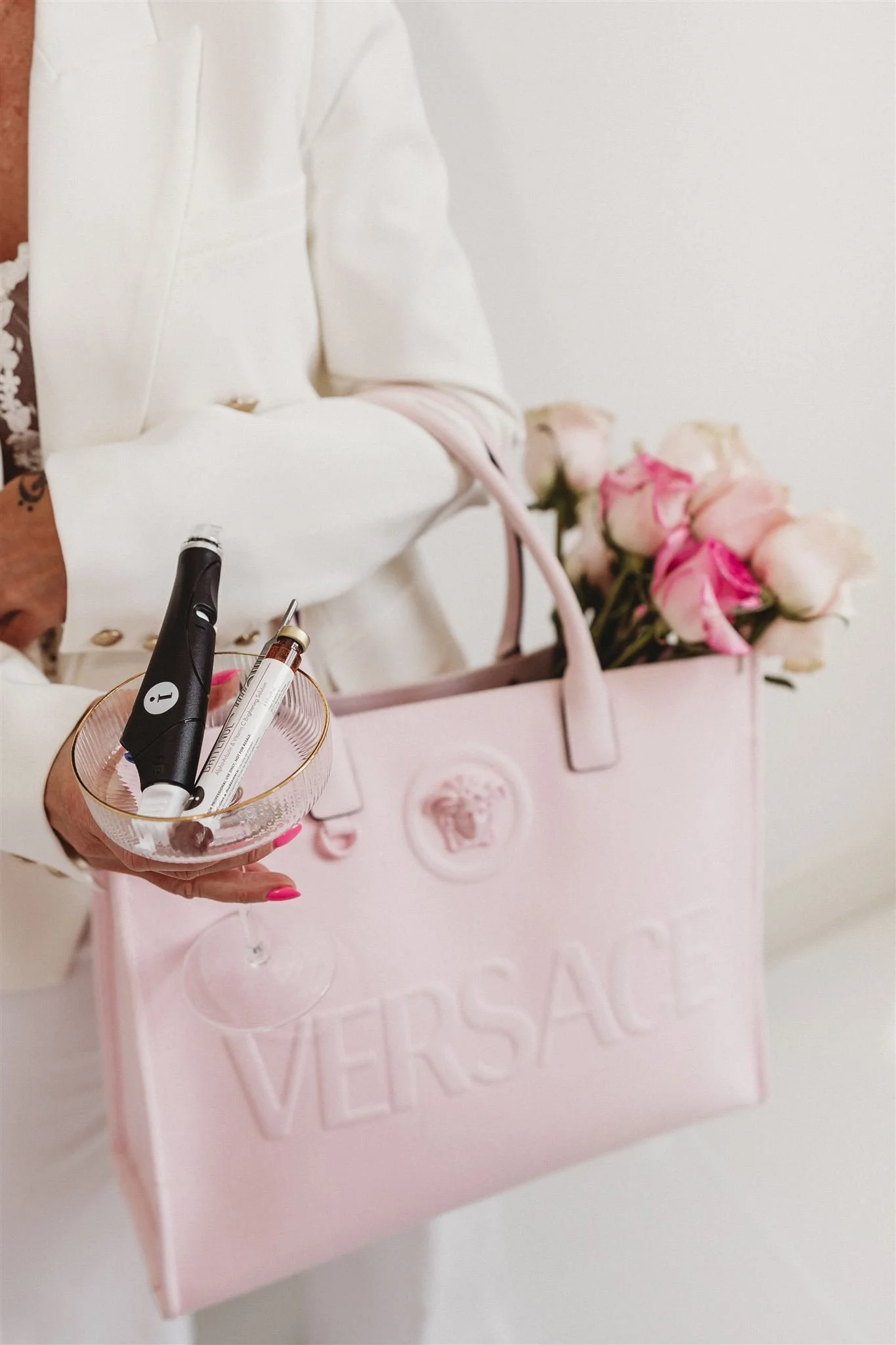 A person holding a pink Versace bag filled with pink and white roses. The person is dressed in white and is holding a glass dish with a tube, a pen, and a small electronic device.