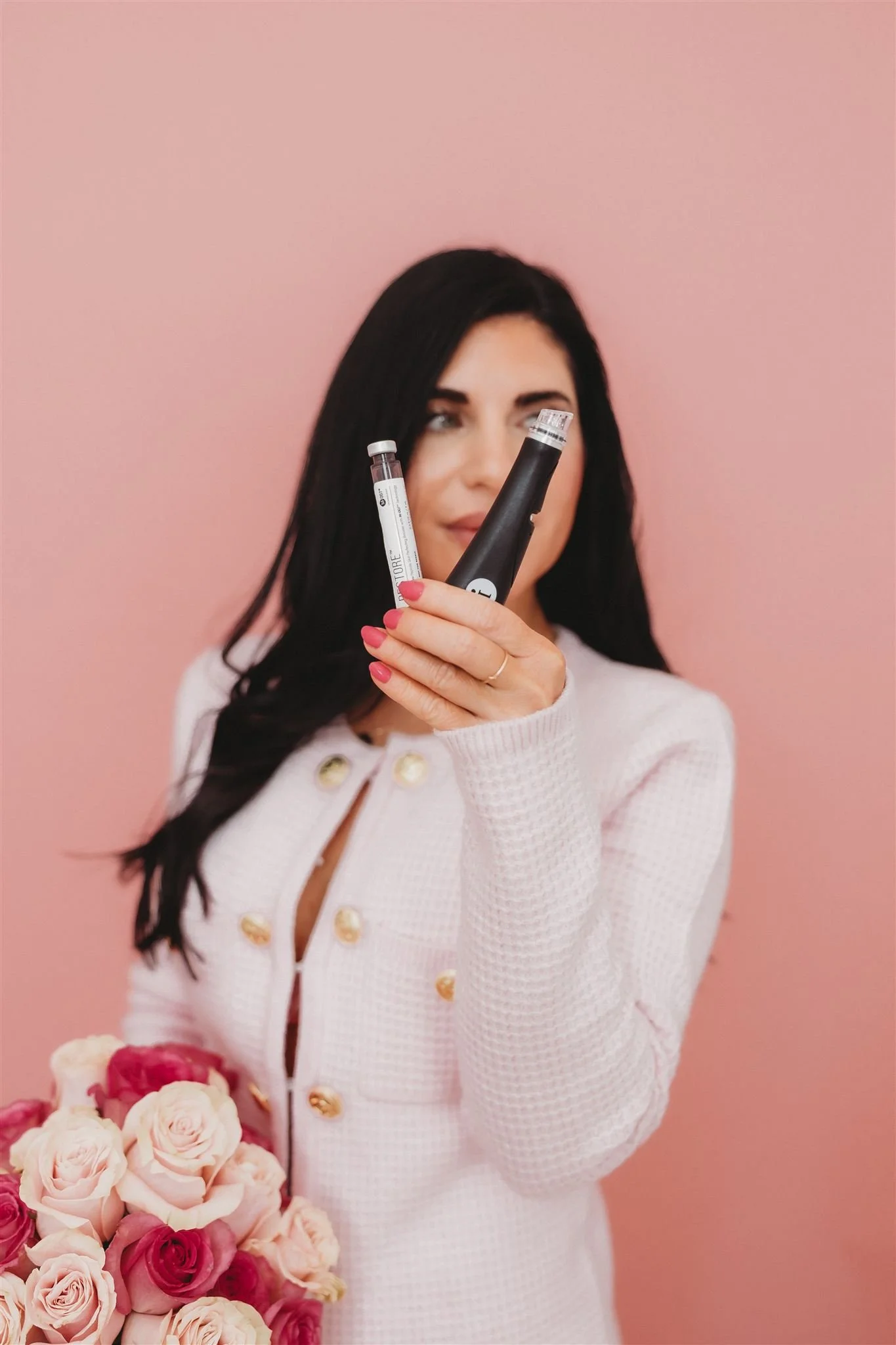A woman with dark hair holding a white and black electronic device in front of a pink background, holding a bouquet of pink roses.