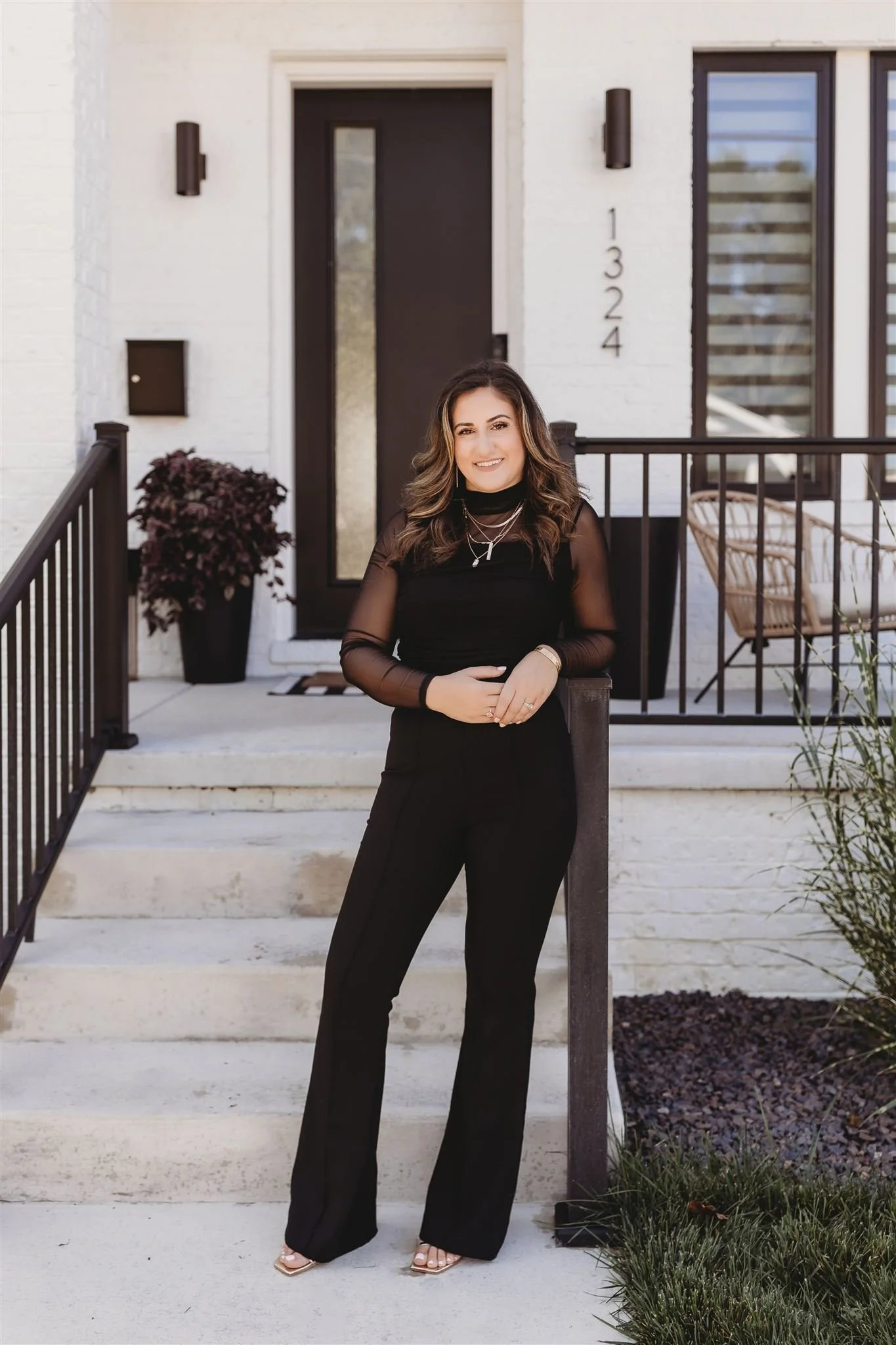 A woman in black clothing posing on front steps of a white modern house with black door and window accents, with house numbers 1324 visible.