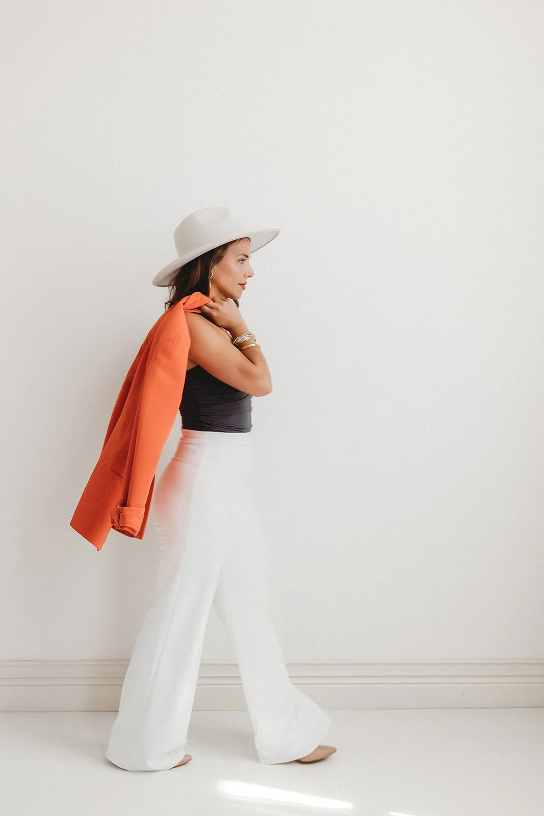 A woman in white wide-leg pants, a black sleeveless top, a white hat, and an orange blazer draped over her shoulder, standing sideways against a plain white wall.