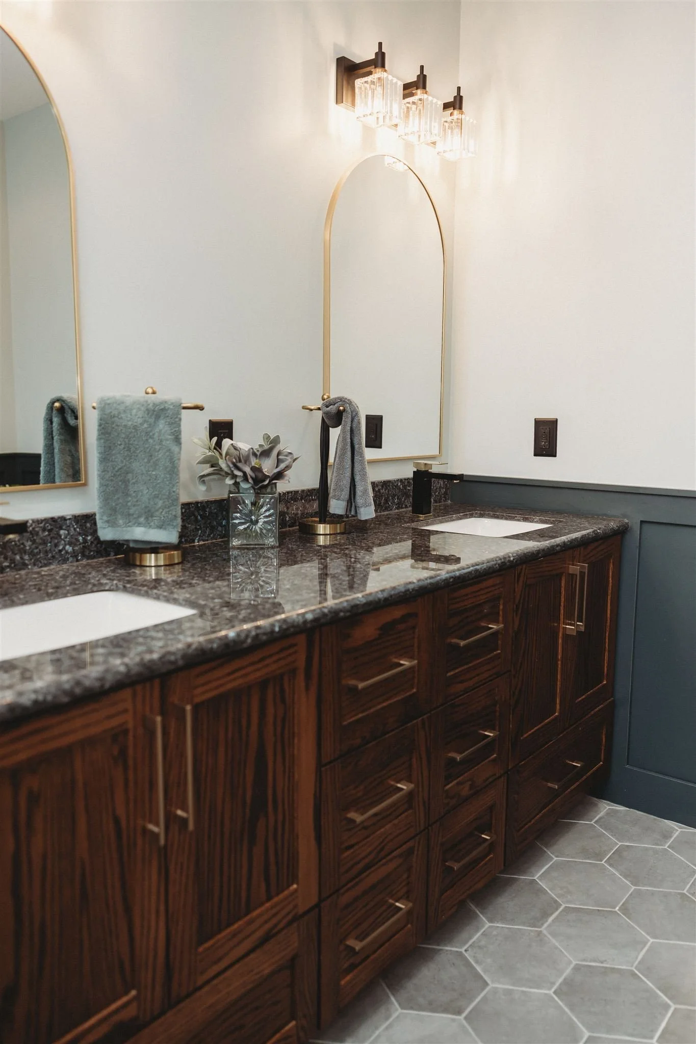 A bathroom vanity with a dark granite countertop, two white sinks, and wooden cabinets with sleek handles. There are two mirrors above the sinks, a modern light fixture with multiple glass shades mounted on the wall, and a towel rack with a green towel and a gray towel. A small potted plant is placed between the mirrors, and the floor is covered with hexagonal gray tiles.