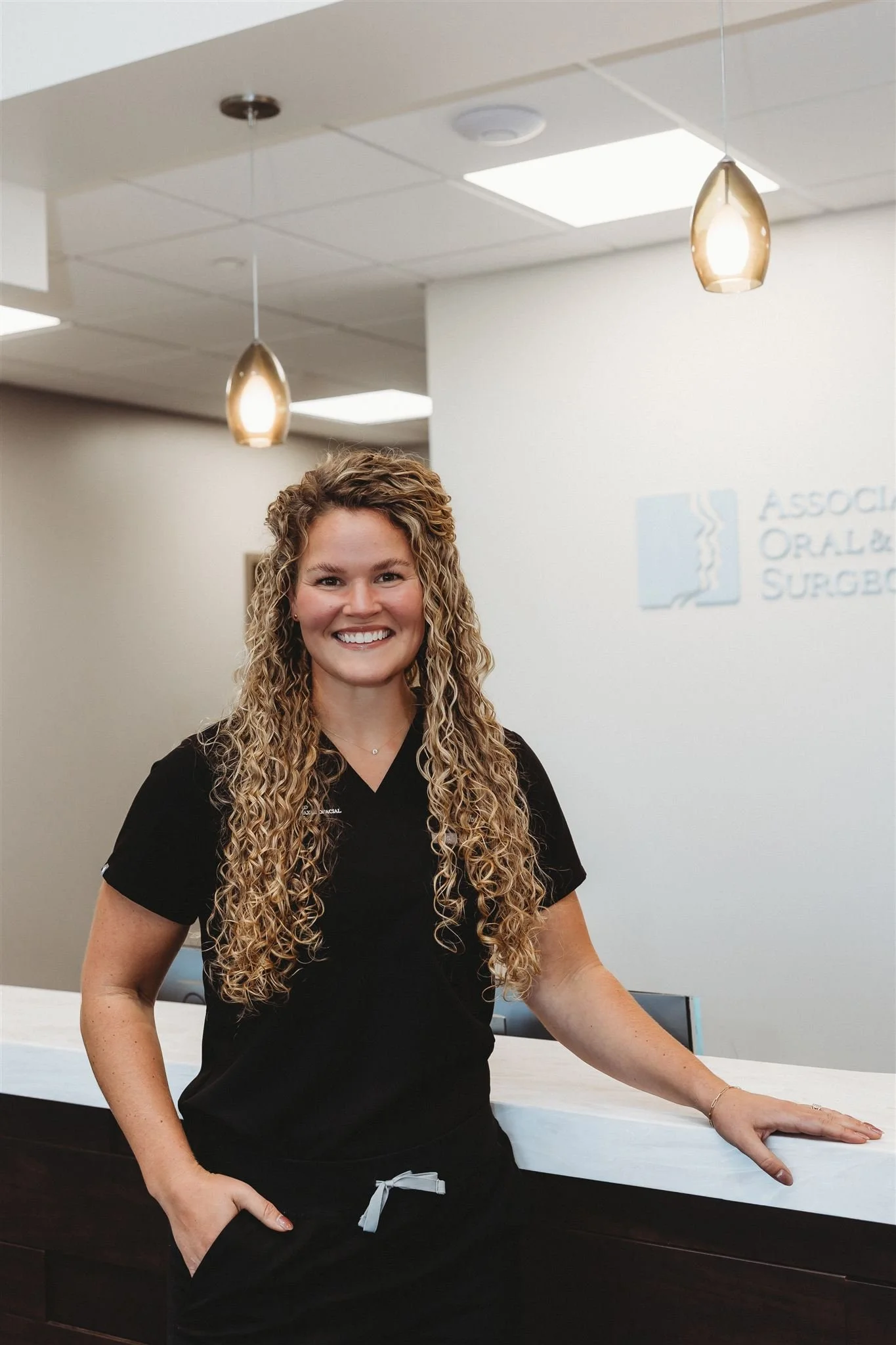 A woman in black scrubs with curly long hair standing at a reception desk in a medical office.