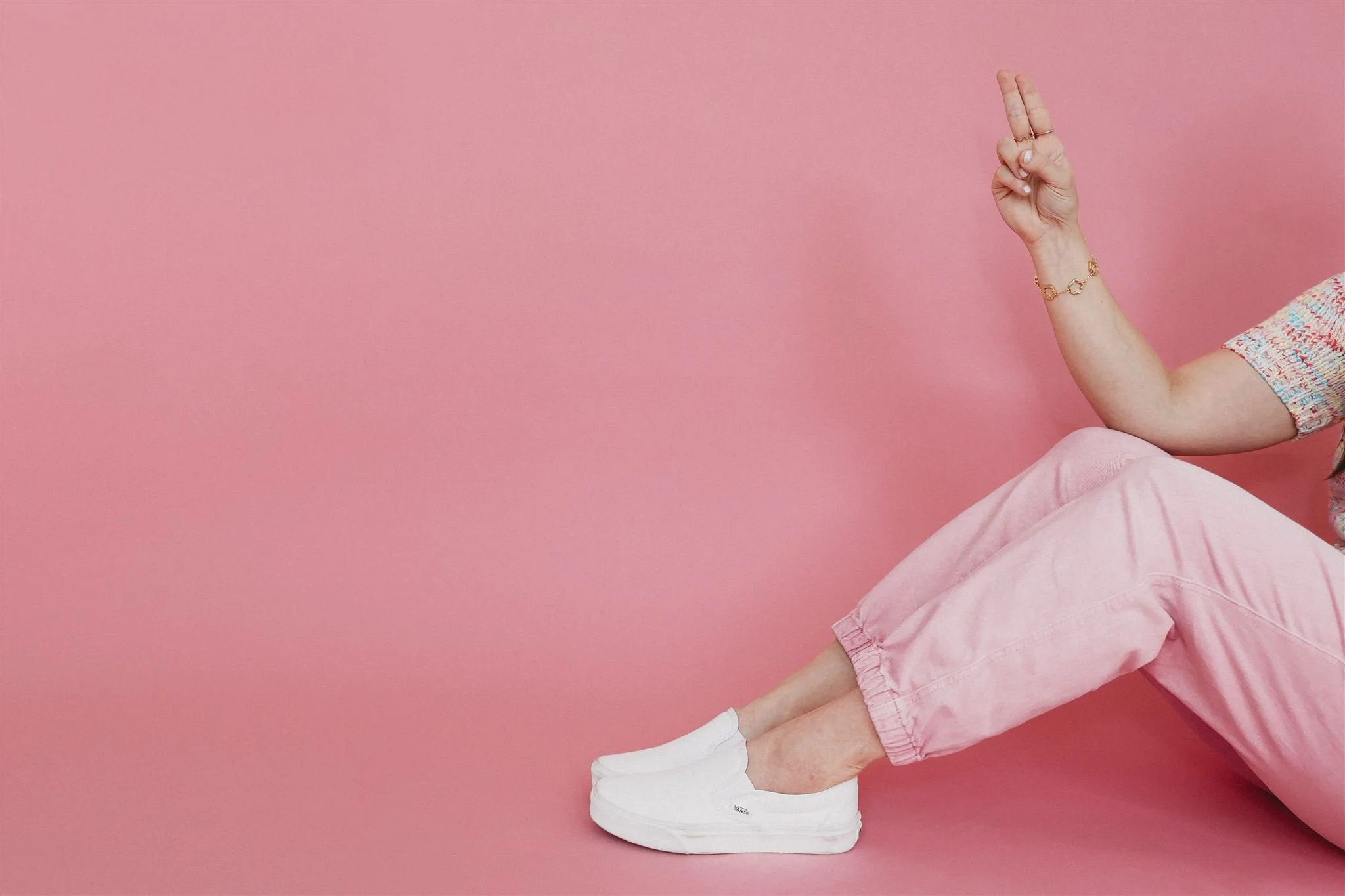 A woman sitting against a pink background wearing pink pants and white Vans slip-on sneakers, with one arm raised showing a peace sign, her face out of frame.
