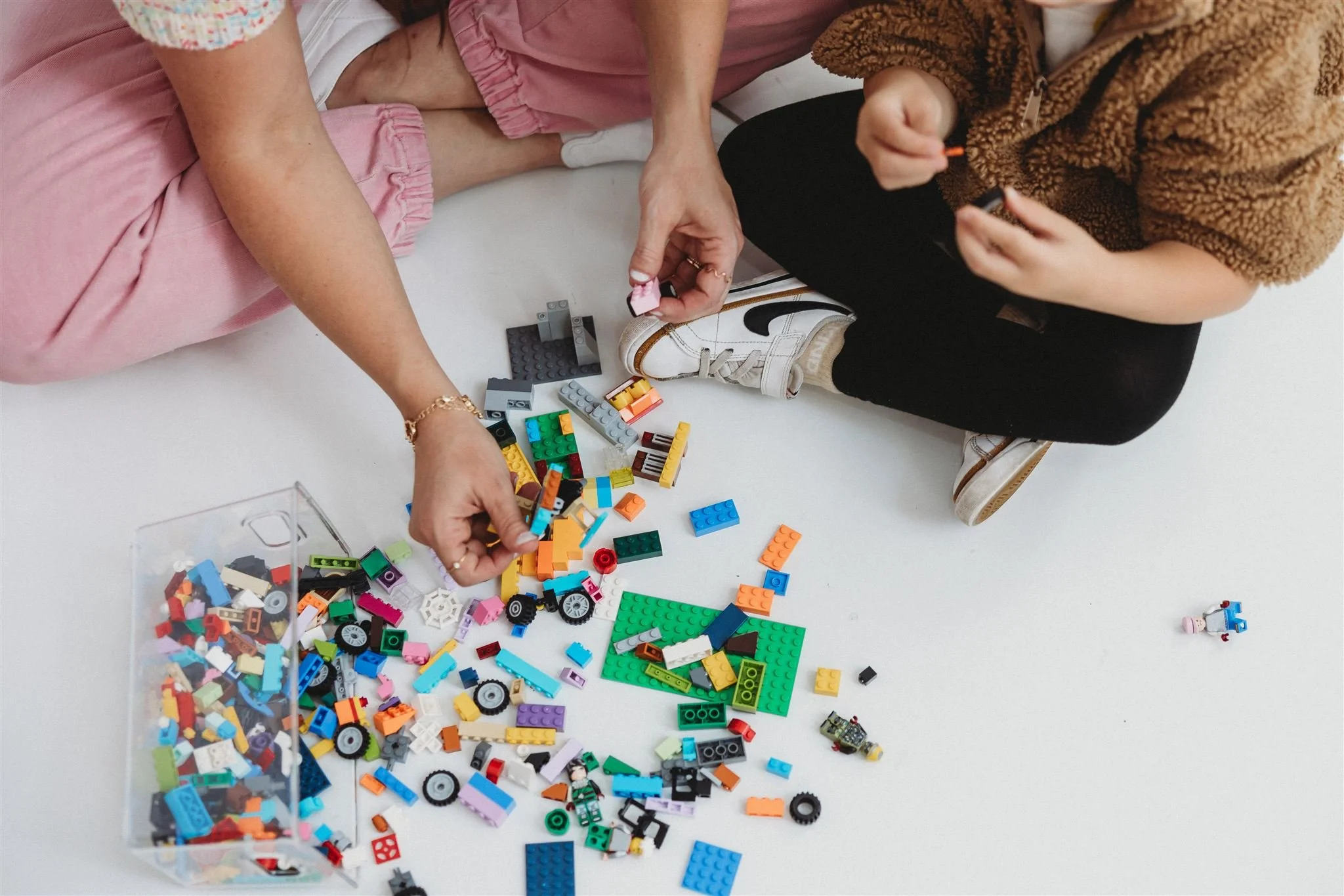 Overhead view of two people sitting on a white floor sorting through a large pile of colorful LEGO bricks spilled from a clear storage container, with small partially built figures nearby.