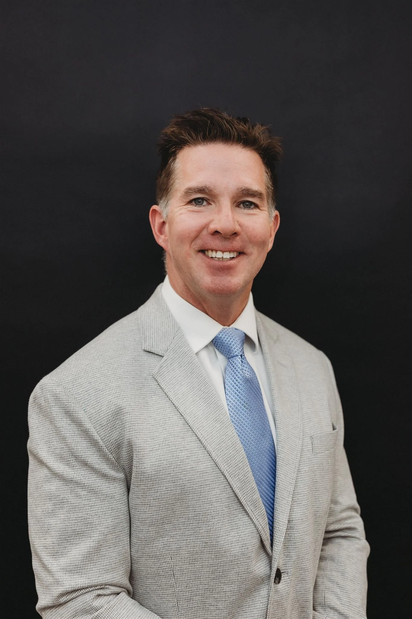 A smiling man in a light gray suit, white shirt, and blue tie, standing against a black background
