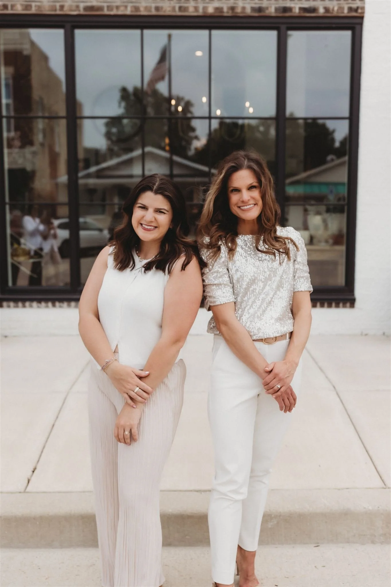 Two women smiling and standing outdoors in front of a large window, dressed in light-colored clothing.