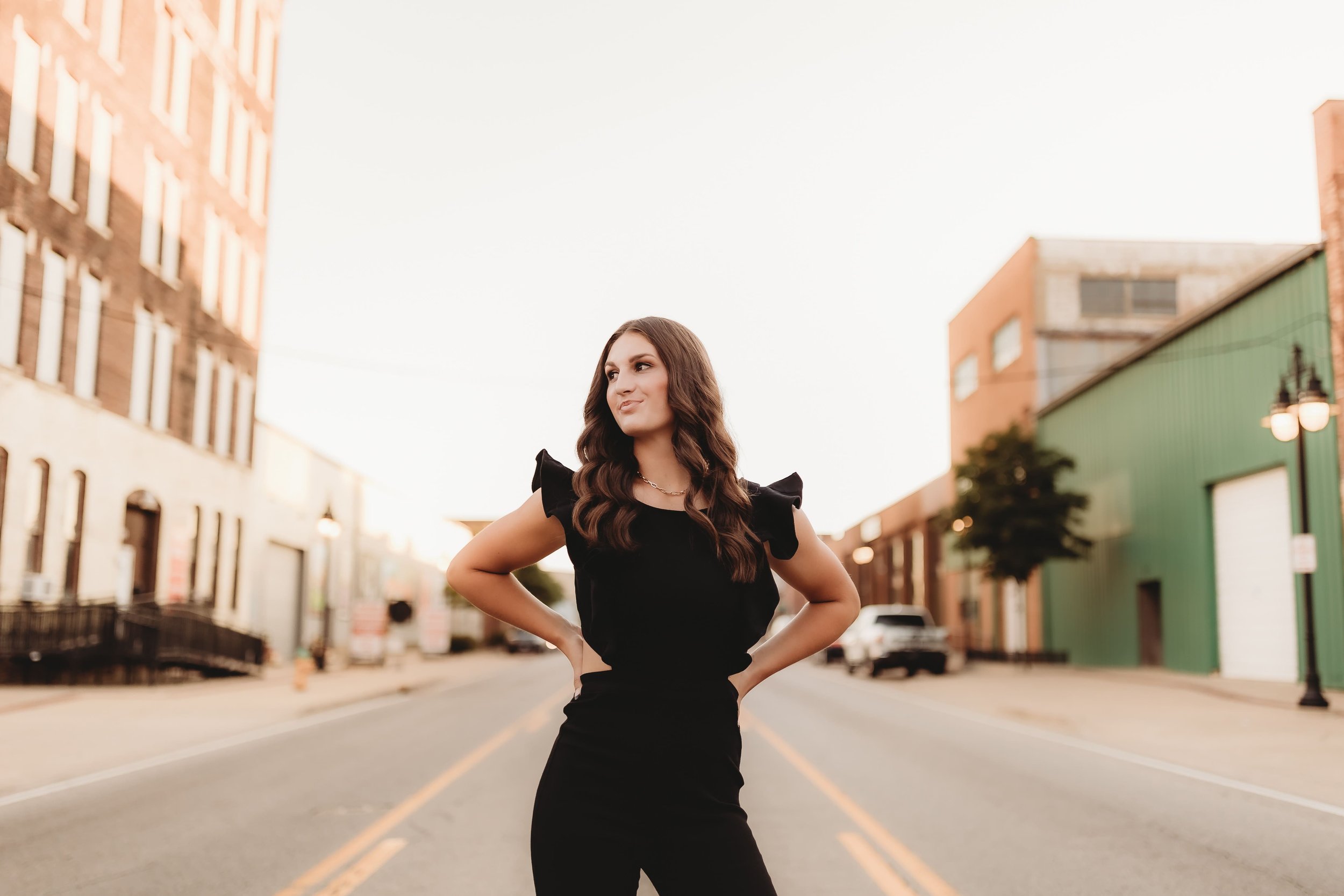  High school senior stands in the middle of an unbusy street in small town illinois with her hands on her hips and looks off into the distance during a photoshoot for senior picture ideas 