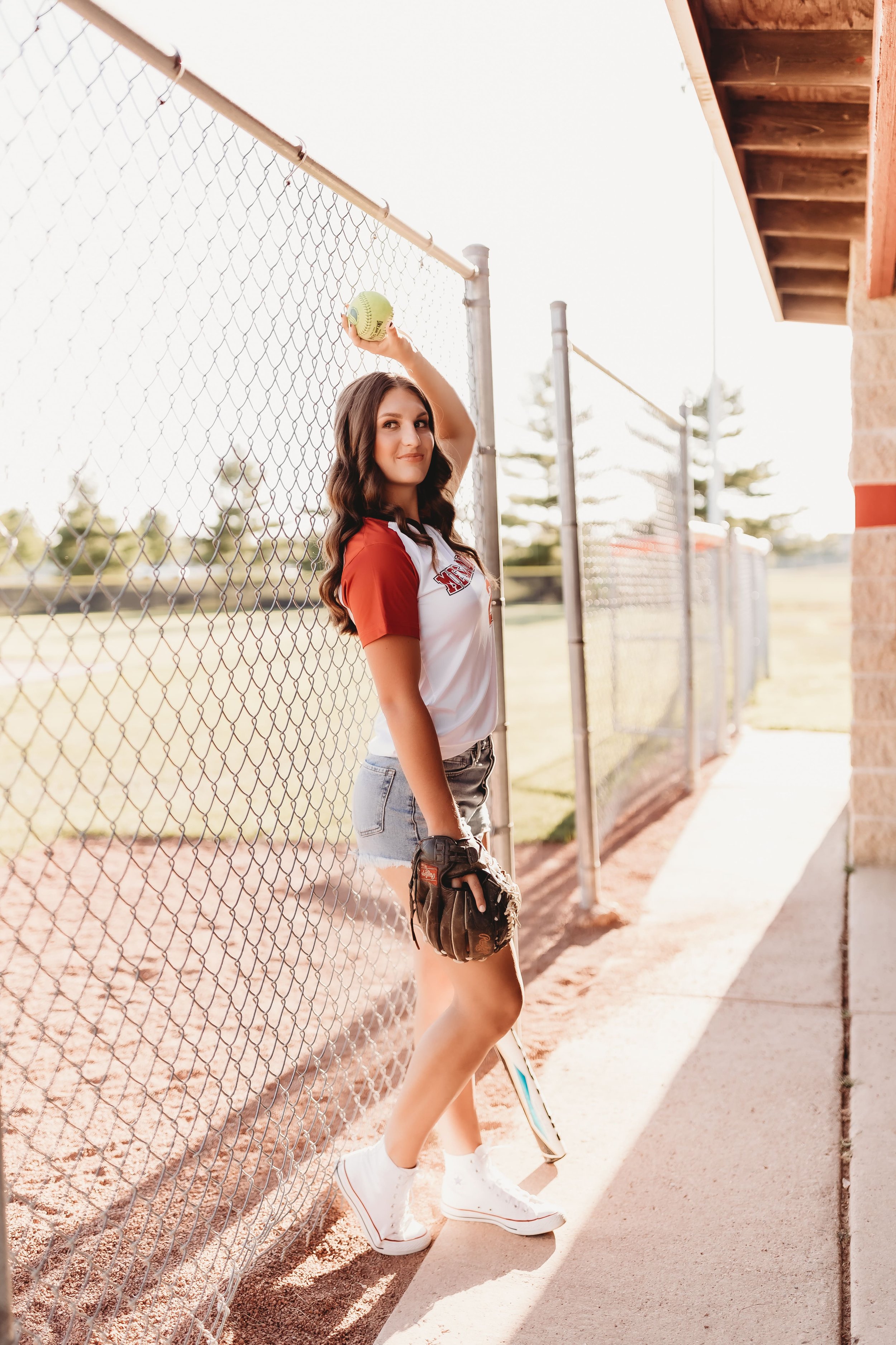  High school senior leans against the chain link fence of a dugout while holding a softball in one hand with a softball glove and smiles while testing softball senior picture ideas in metamora Illinois 