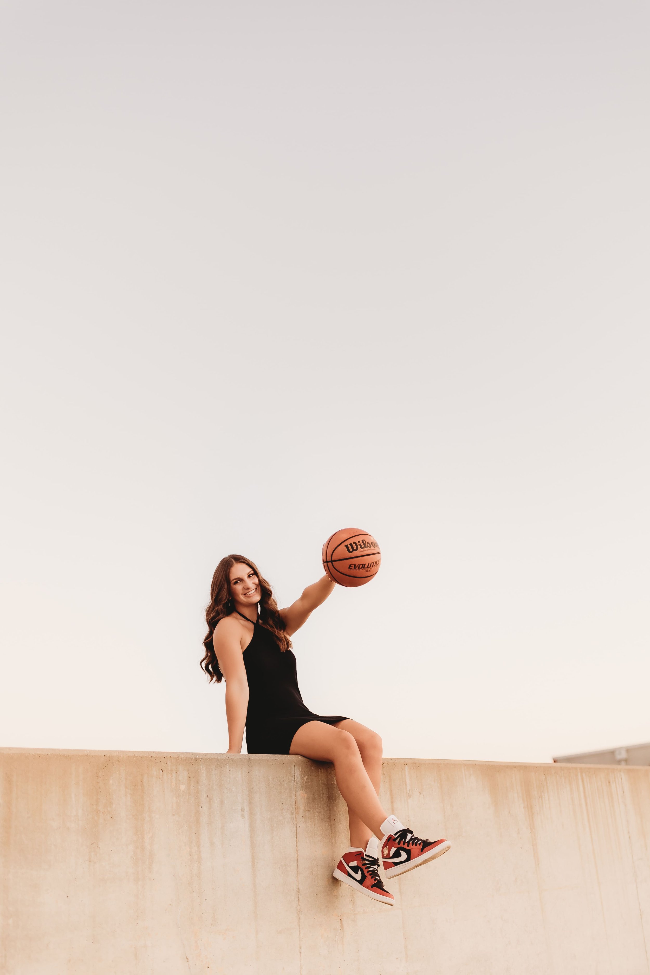  High school senior smiles as she sits on the edge of a rooftop and palms a basketball while taking senior pictures basketball 