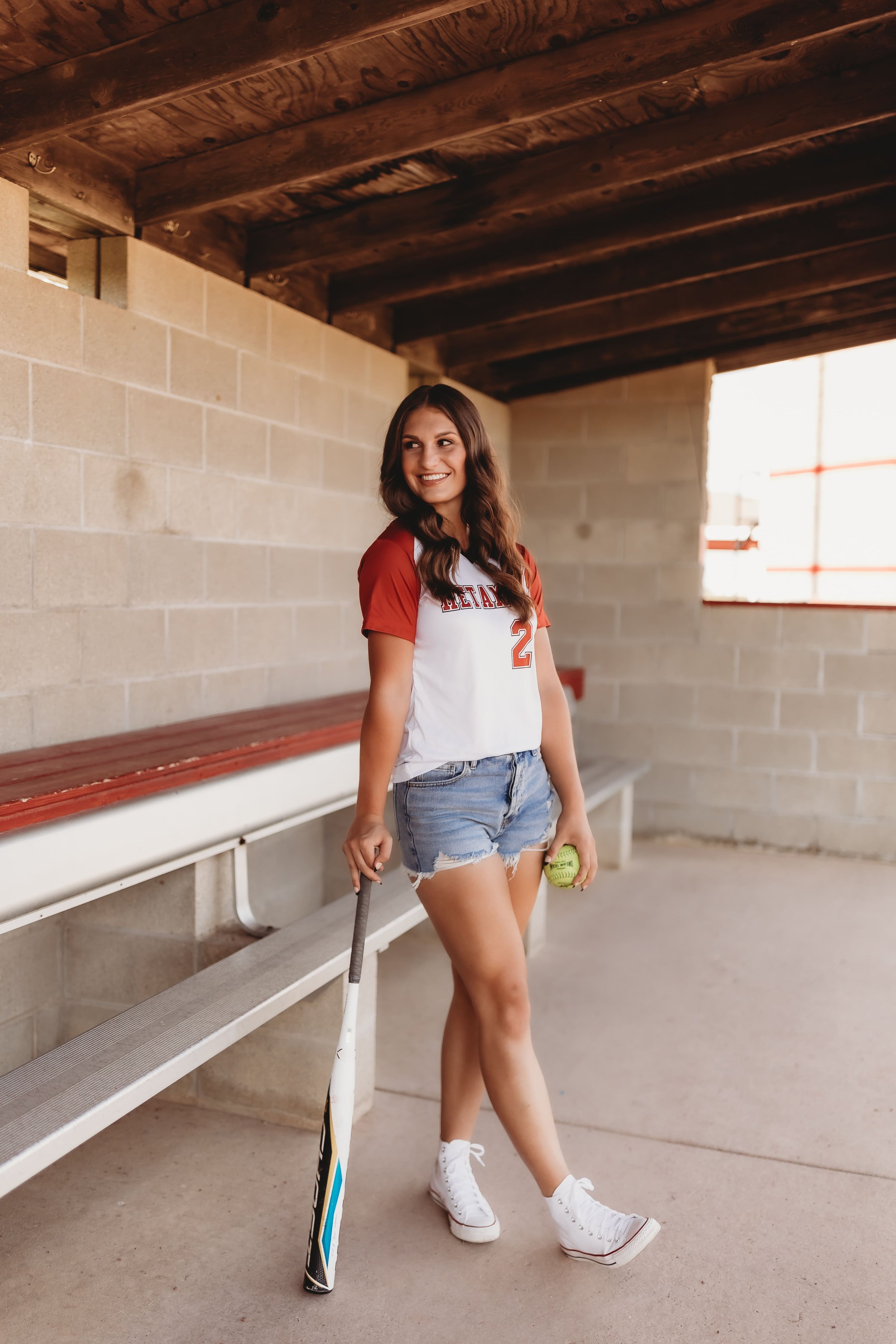  Senior softball player stands in a dugout and leans on her softball bat and smiles over her shoulder as she holds a softball senior picture ideas 