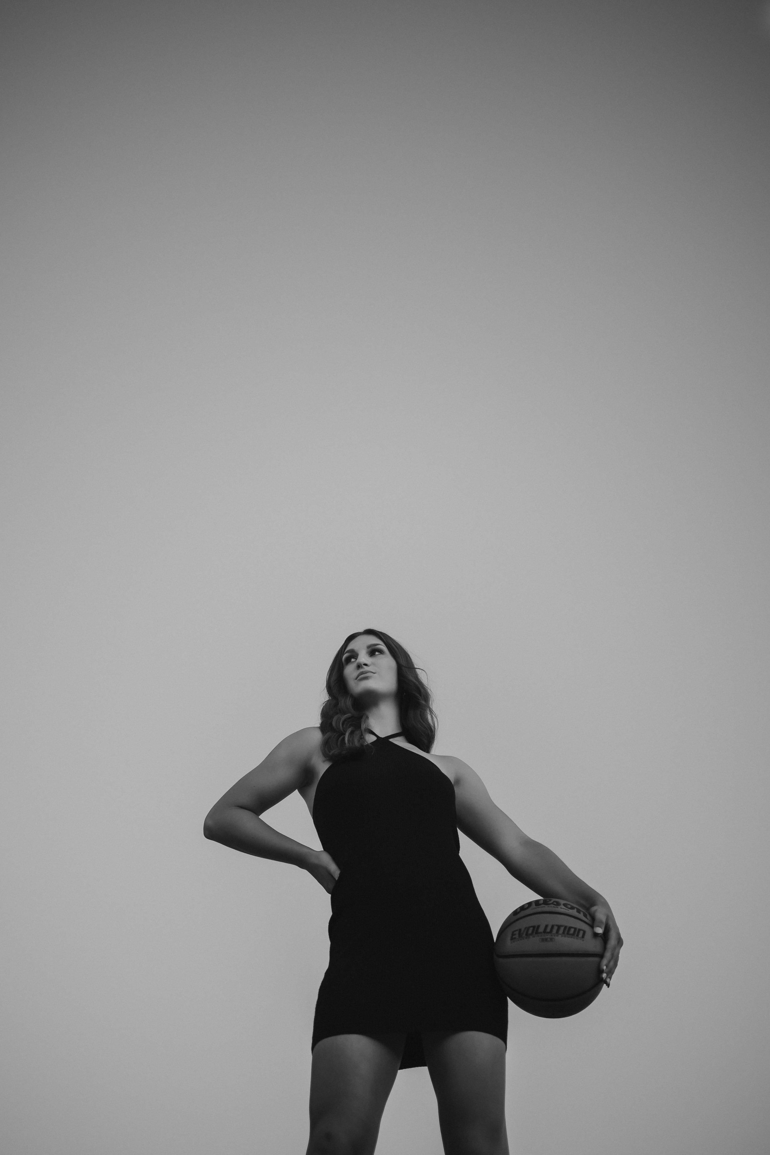  A black and white photo of a high school senior wearing a black dress and holding a basketball shot from a low angle posing for senior pictures basketball 