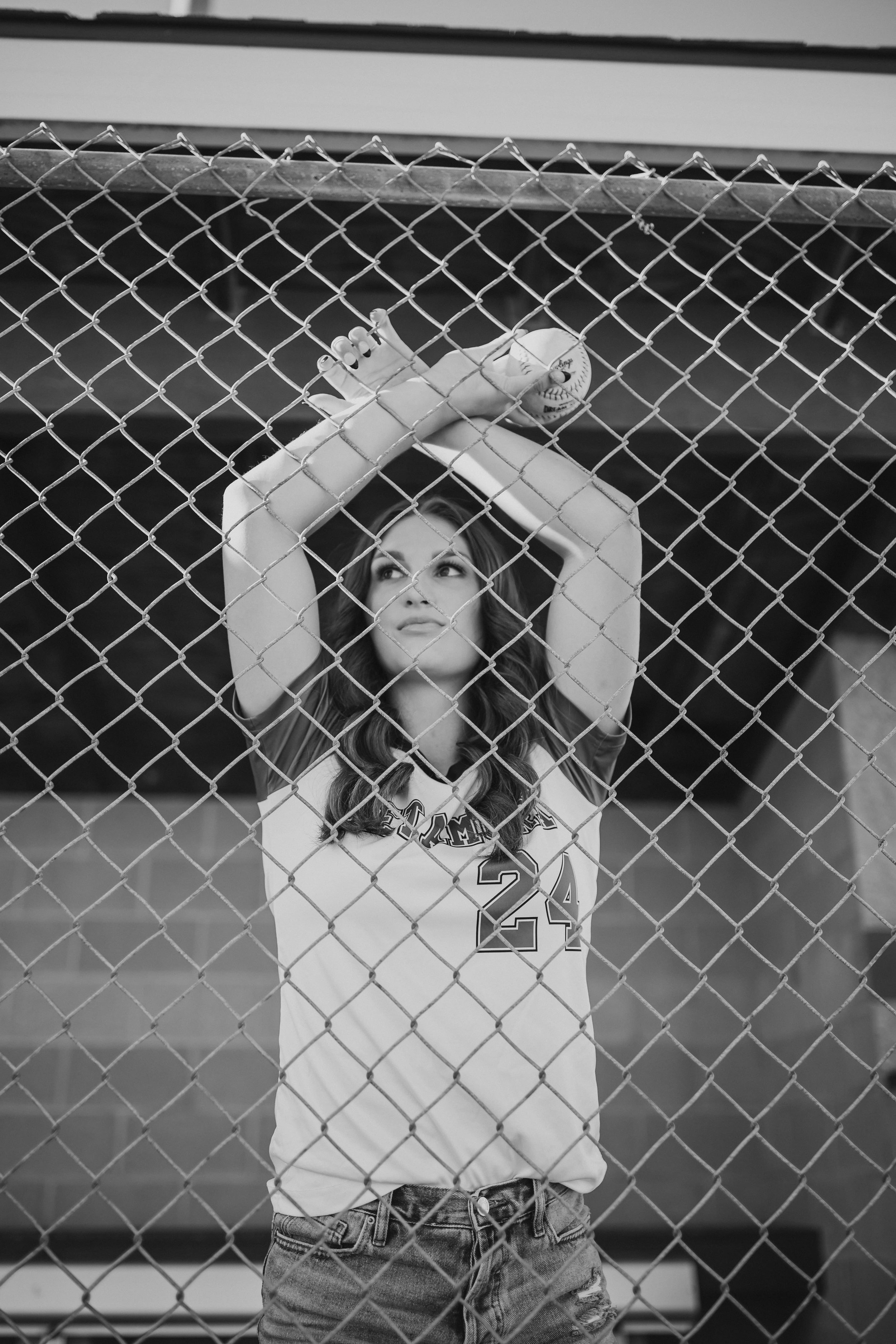  Senior softball player leans forward on the chainlink fence of a dugout and looks off into the distance as her photographer captures her for softball senior picture ideas 