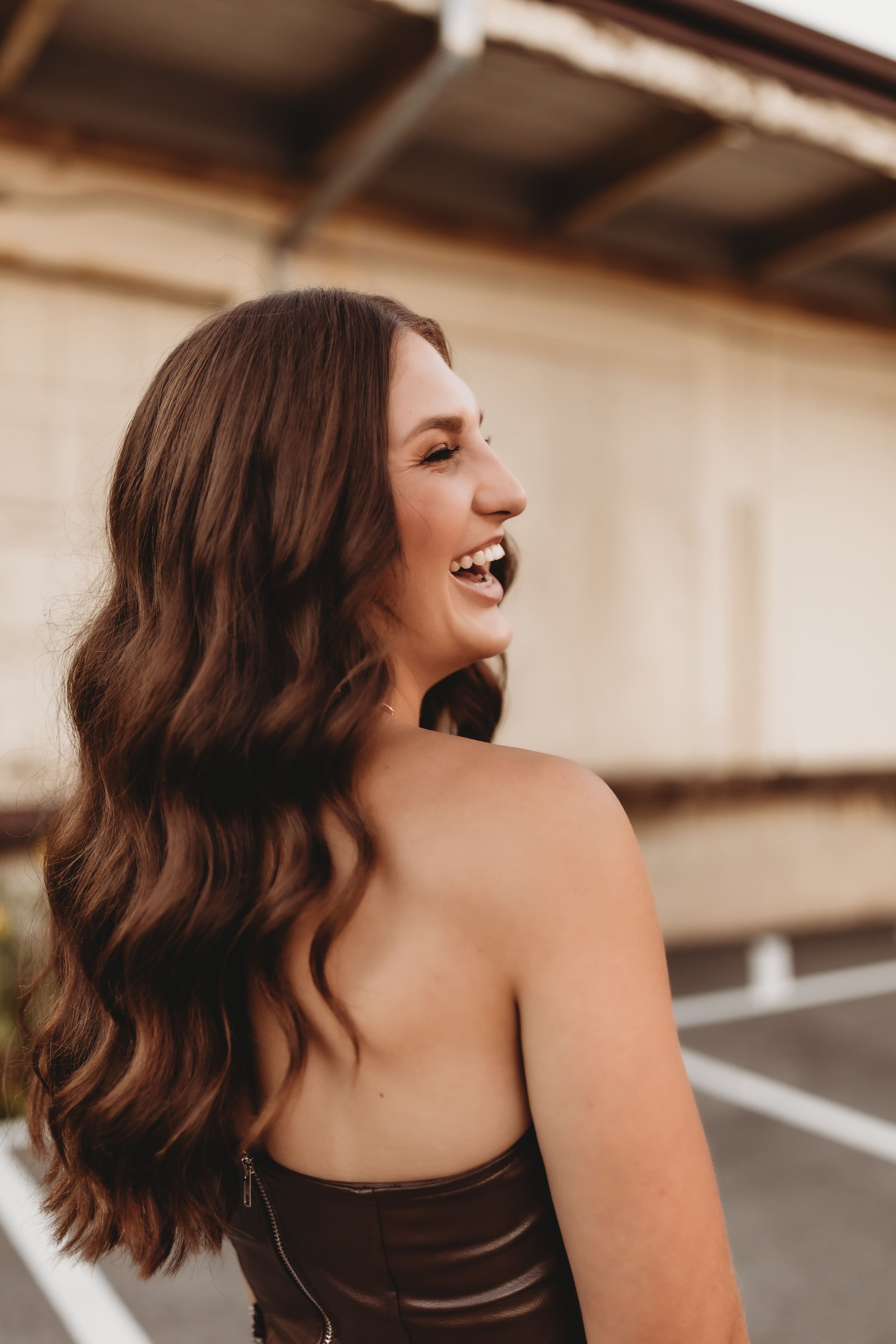  Out of all senior picture photographers Katy chose always flourishing photography seen here laughing with her long brown curls falling over her shoulder as she stands in front of an industrial building 
