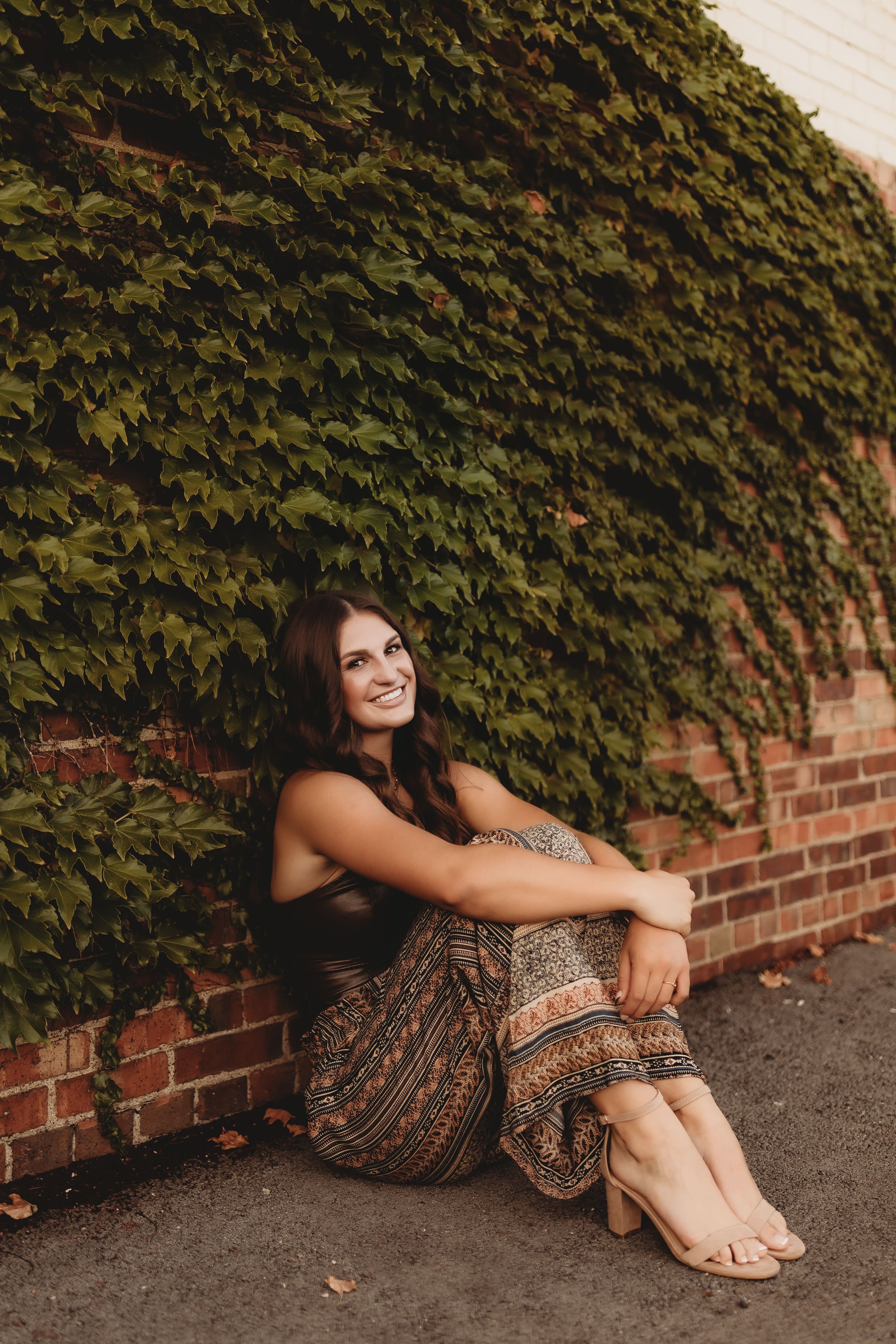  High school senior leans against a brick wall covered in ivy and smiles while holding her knees while posing for senior picture photographers 