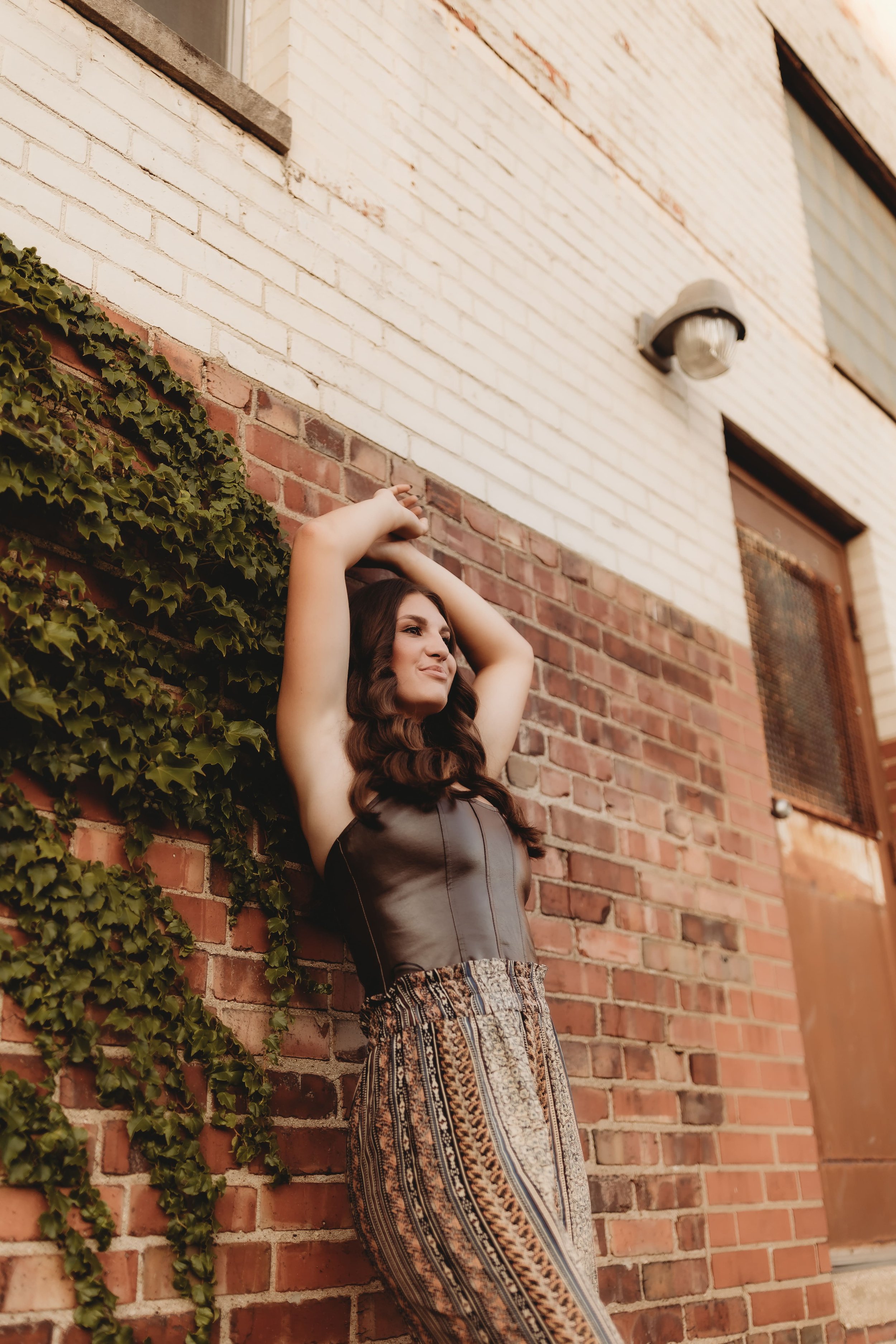  Katy holds her arms over her head while leaning against a brick wall covered in ivy while posing for senior photographers near me 