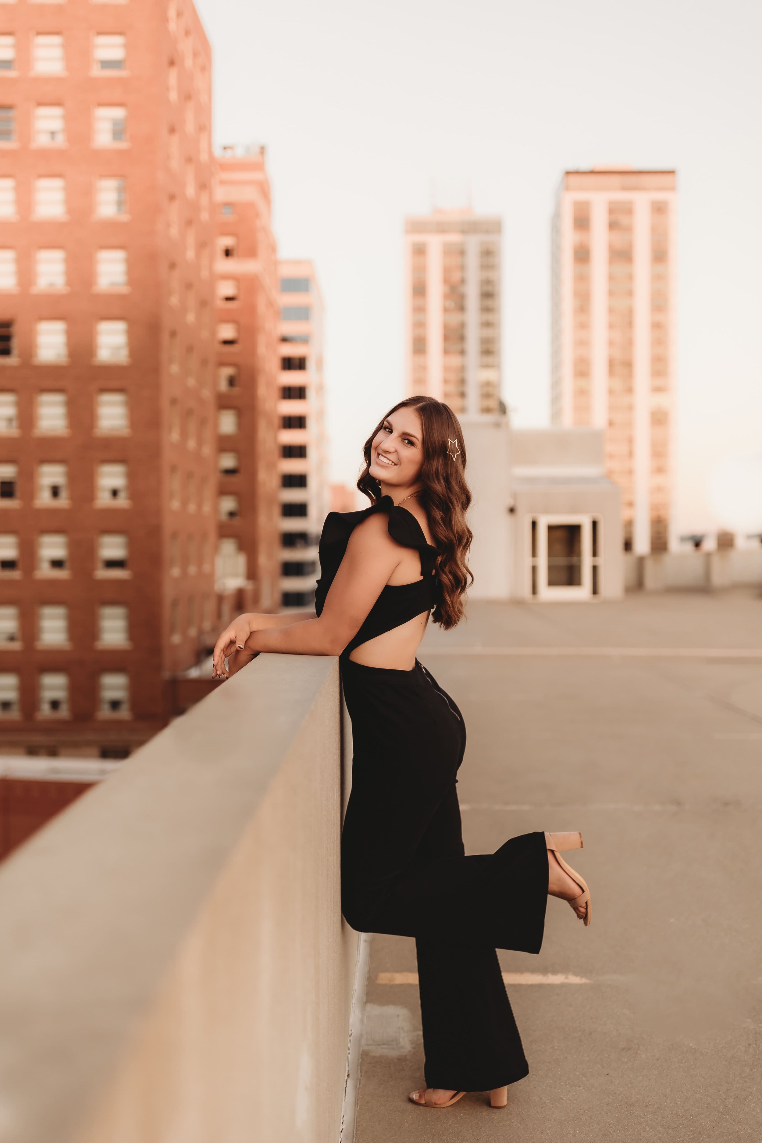  Senior stands near the edge of a downtown peoria rooftop as she smiles over her shoulder with one leg kicked up behind her posing for senior photographers near me 