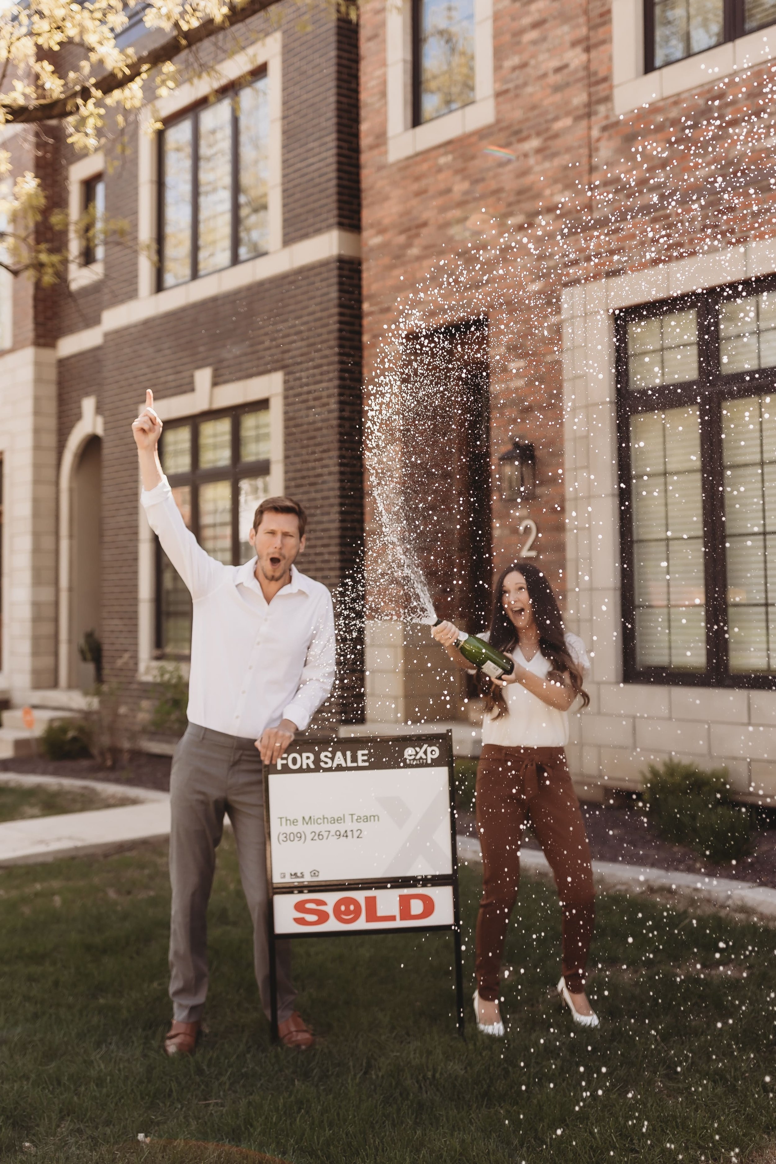  Kayleigh pops a bottle of champagne while cody cheers her on next to their realty for sale sign on the lawn of a peoria brownstone 