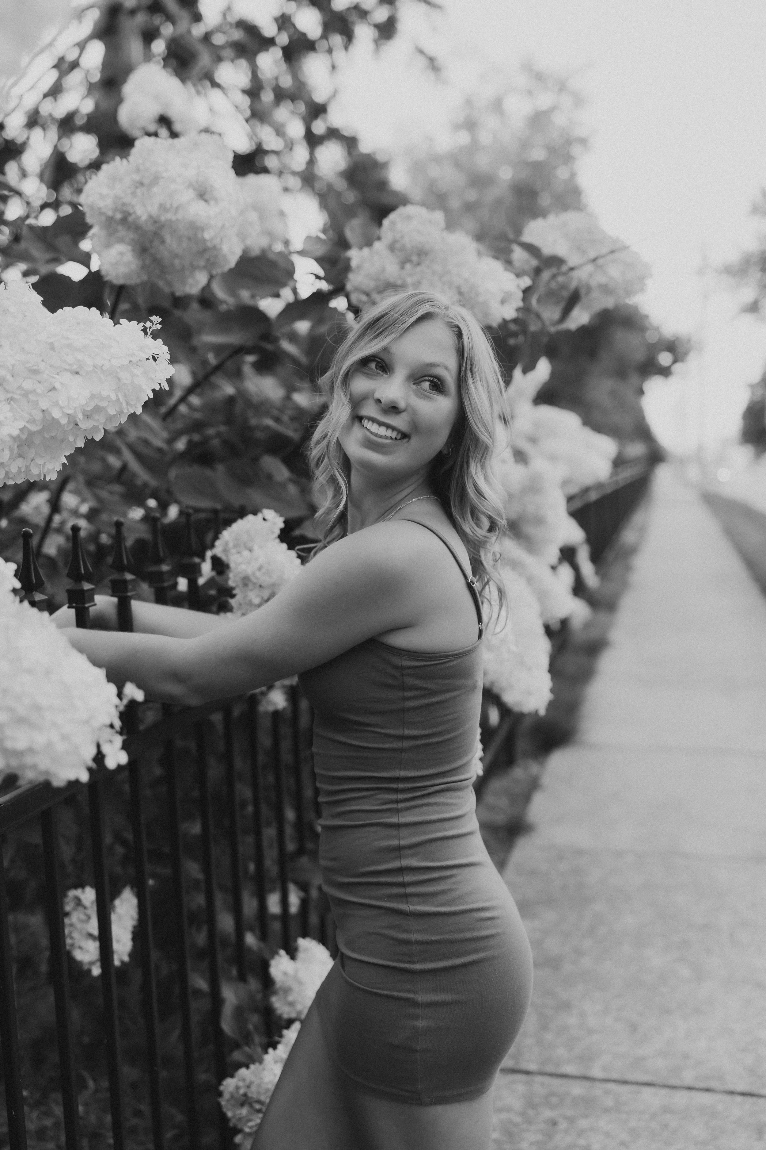  a black and white portrait of a high school senior standing near a flower bush and fence and smiling over her shoulder 