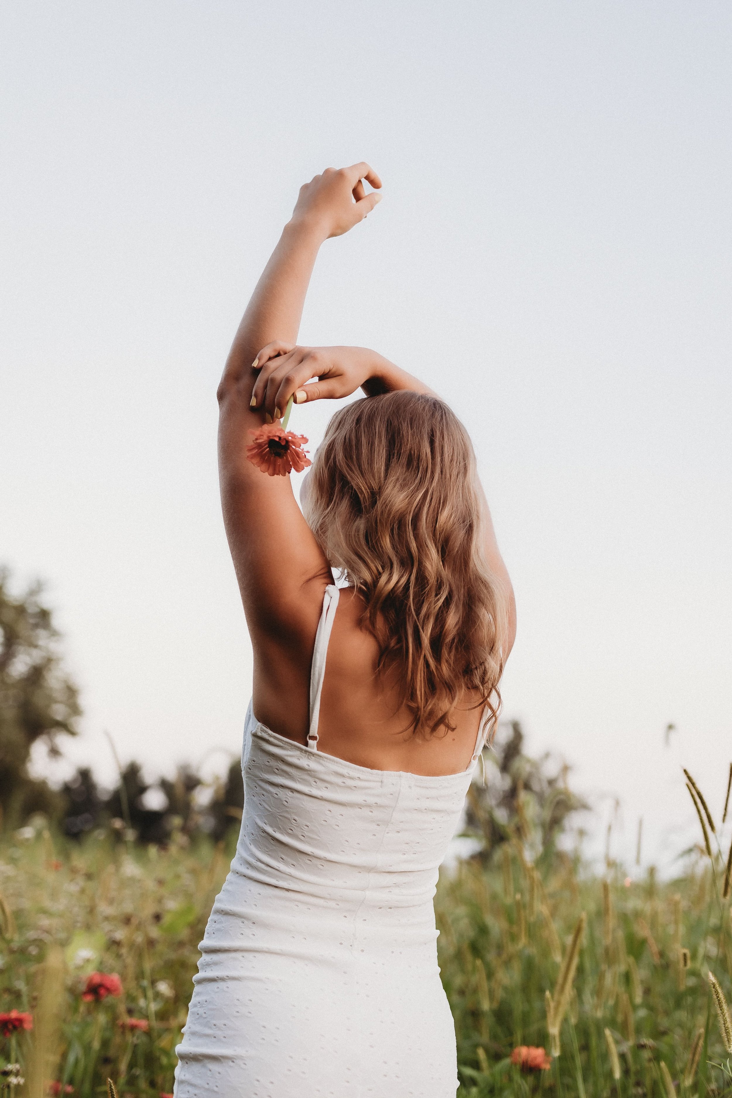  a high school senior stands in a flower field with her arms over her head for senior portraits 