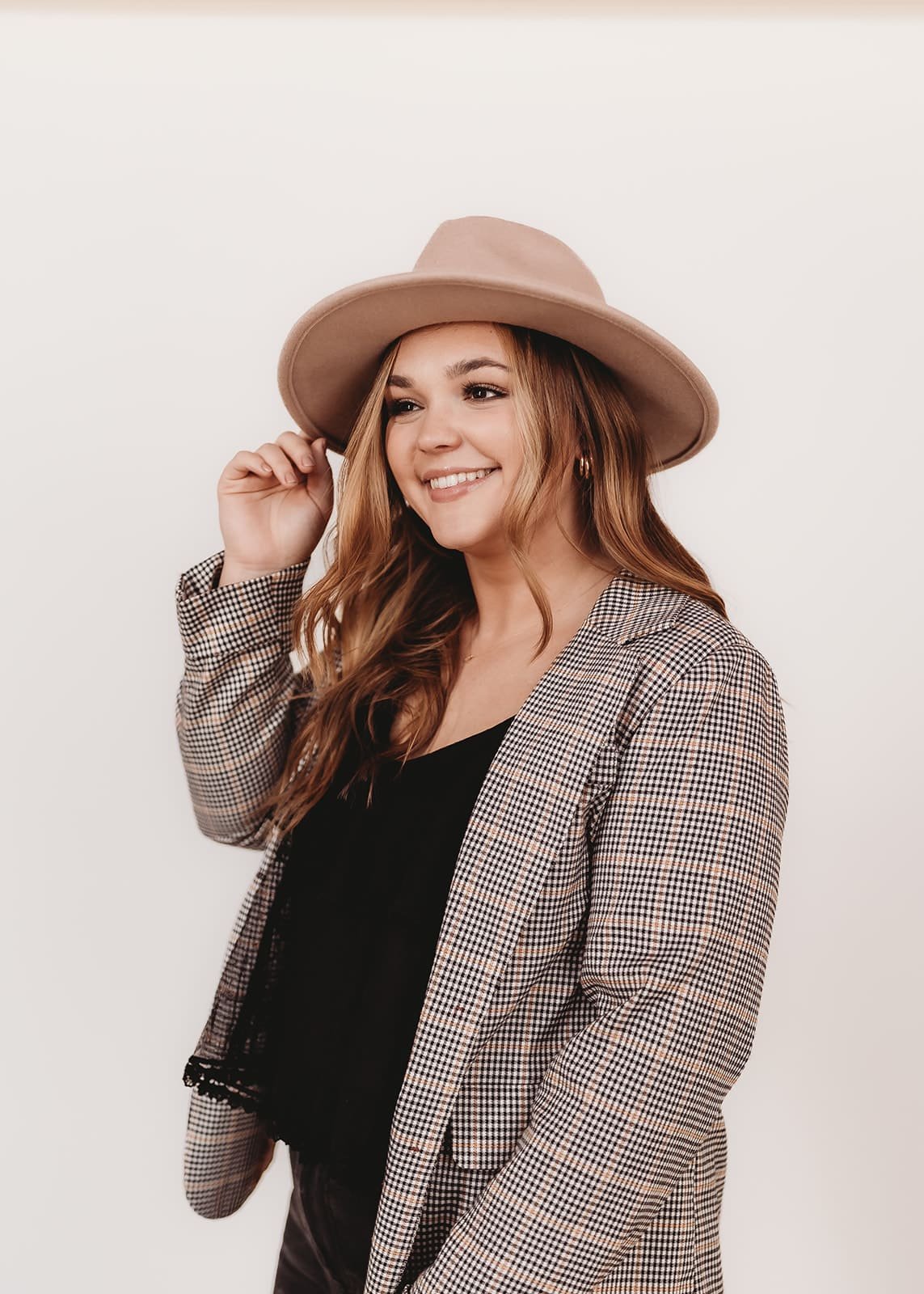  a hair stylist stands in front of a white backdrop while wearing a fun hat and a plaid jacket for a headshot 